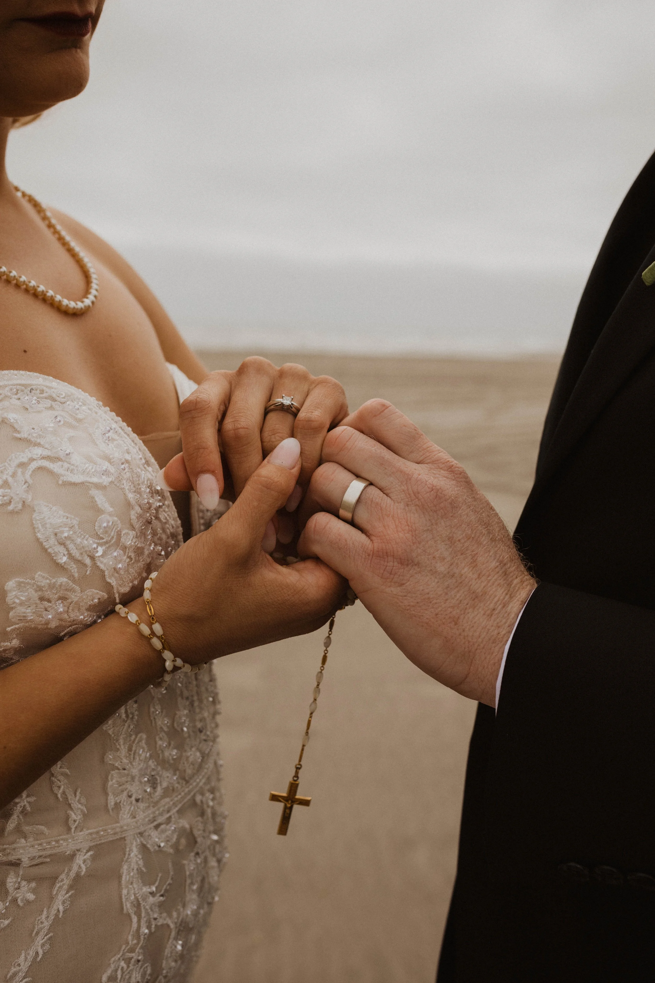 Close-up of a bride and groom holding hands, showcasing their wedding rings, with a beach and ocean in the background. Long Beach, WA wedding photography.