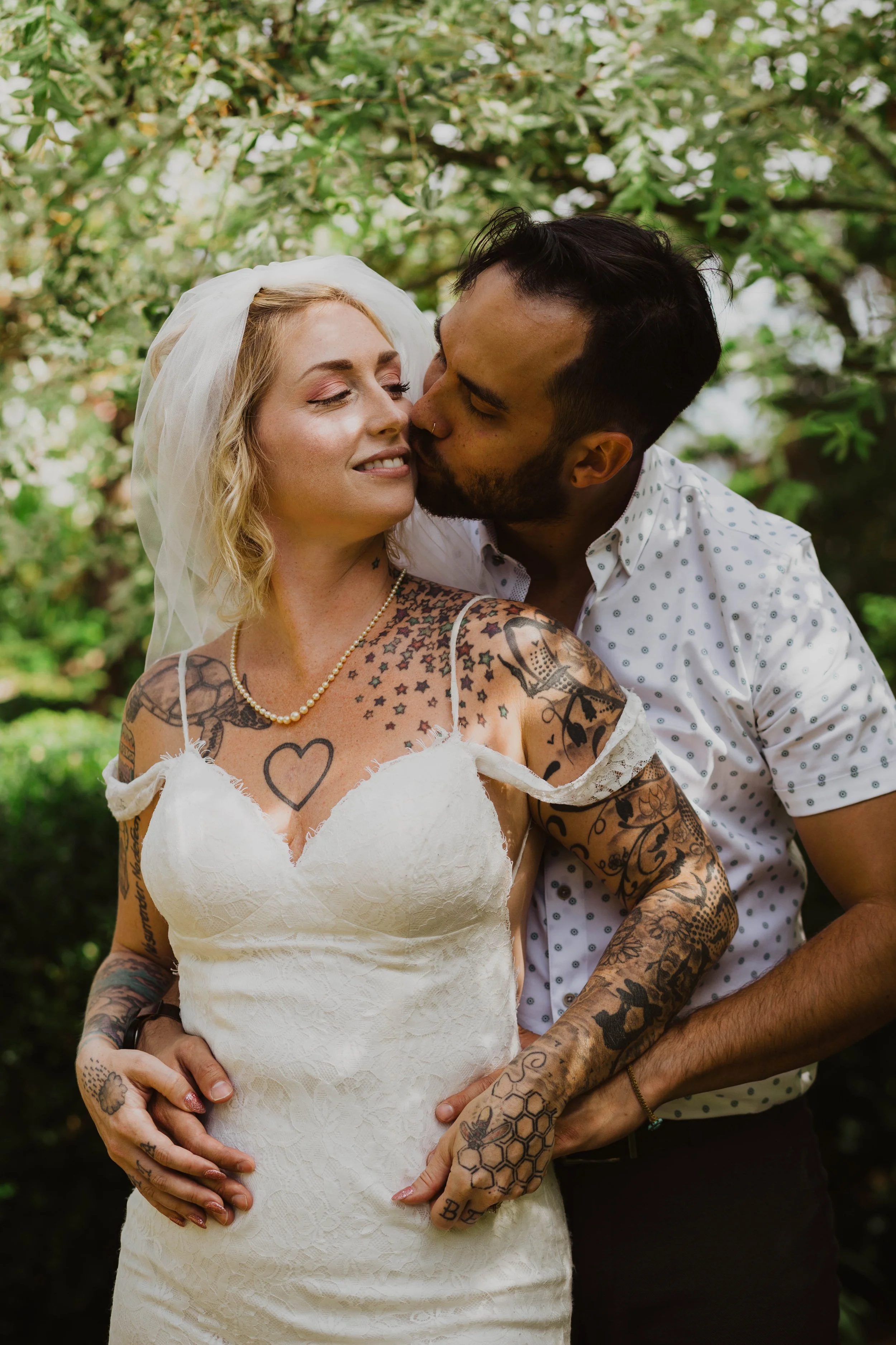 A woman in a white dress and veil being kissed on the cheek by a man in a white polka-dot shirt, both surrounded by green foliage. Seattle, WA wedding photography.