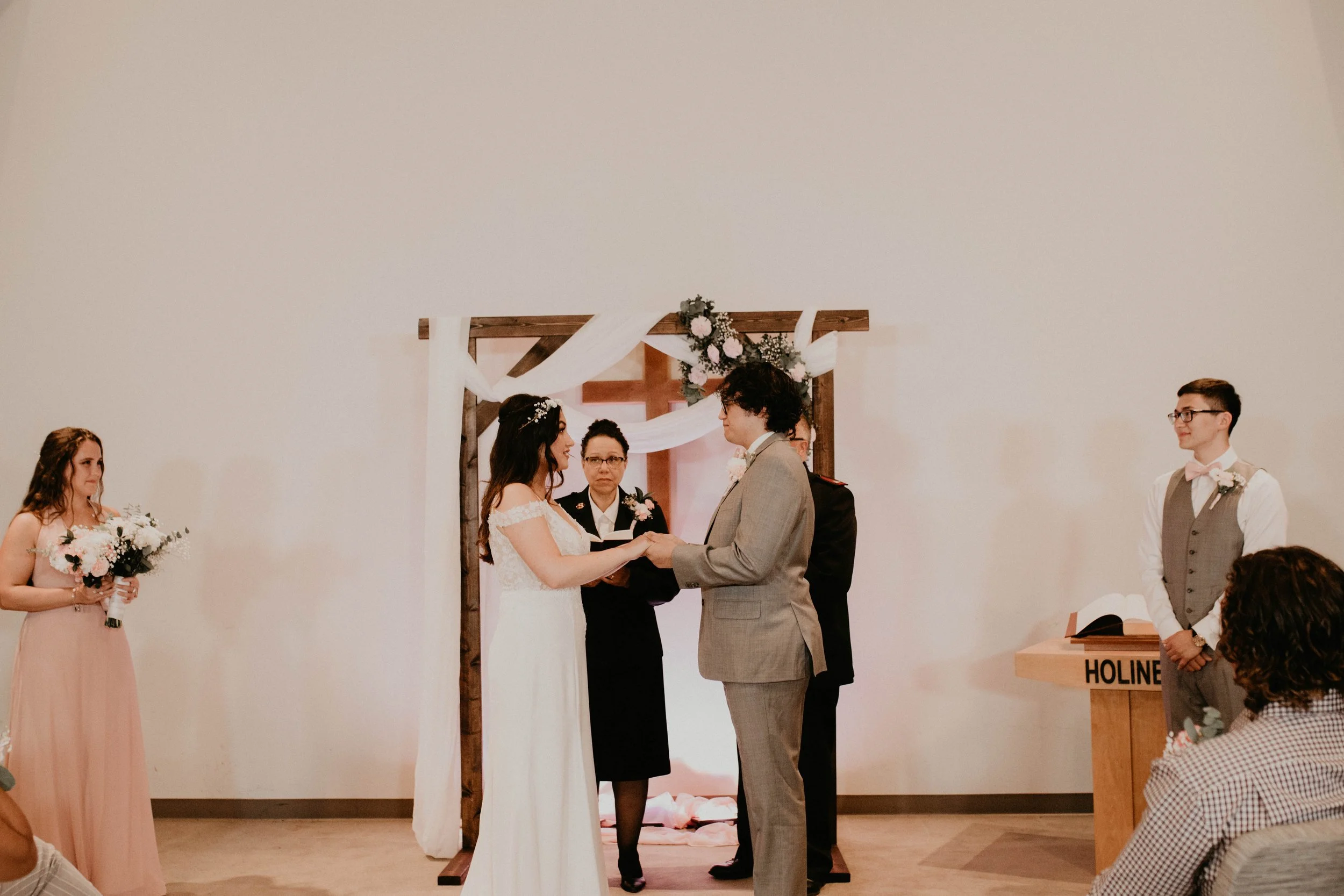 A wedding ceremony with a bride and groom holding hands, with bridesmaids, groomsmen, and an officiant officiating in front of a decorated wooden arch with flowers and white fabric. Seattle, WA wedding photography.