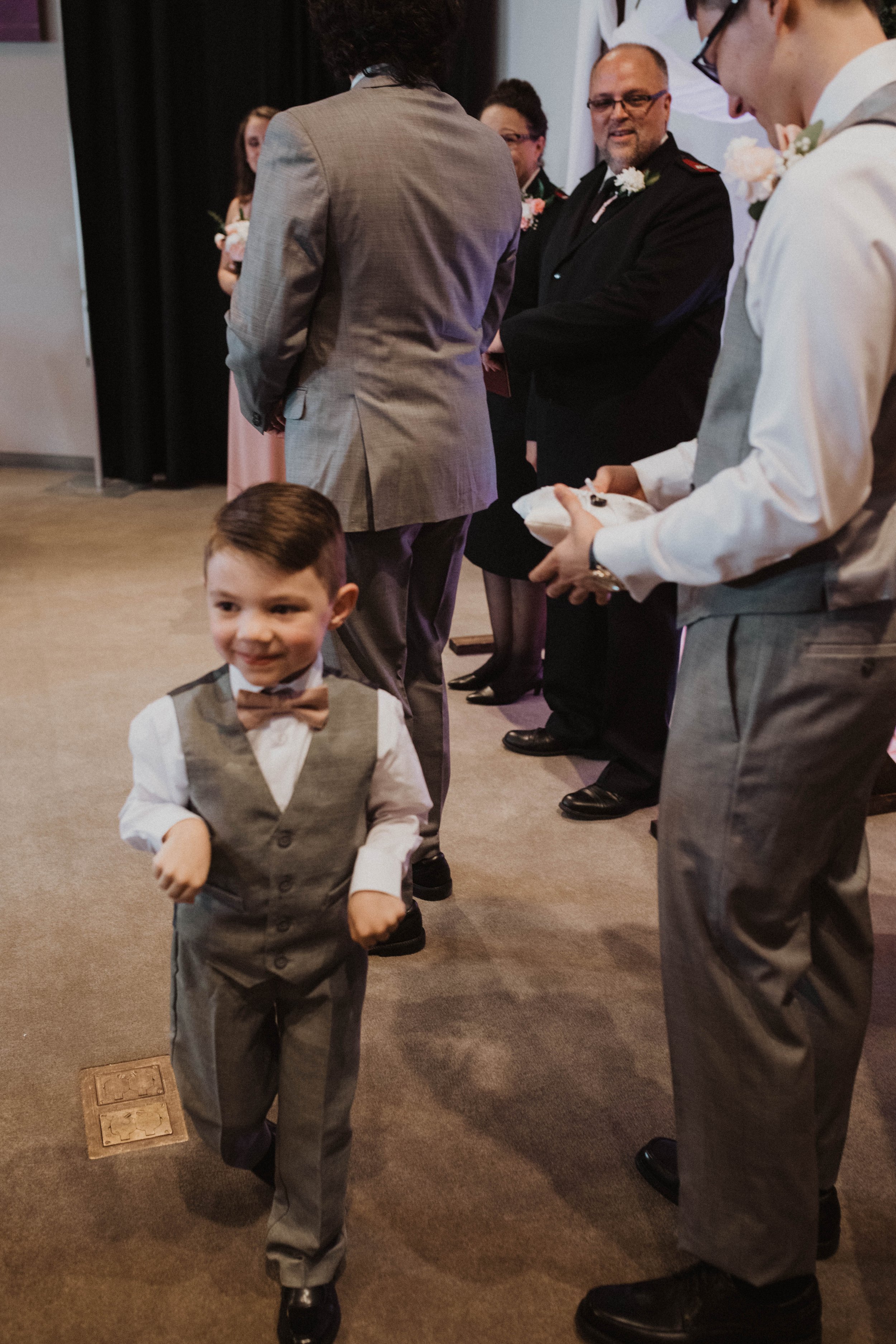 A young boy dressed in a gray vest, white shirt, and bow tie smiling and walking towards the camera at a formal event, with several adults in suits standing in line behind him. Seattle, WA wedding photography.