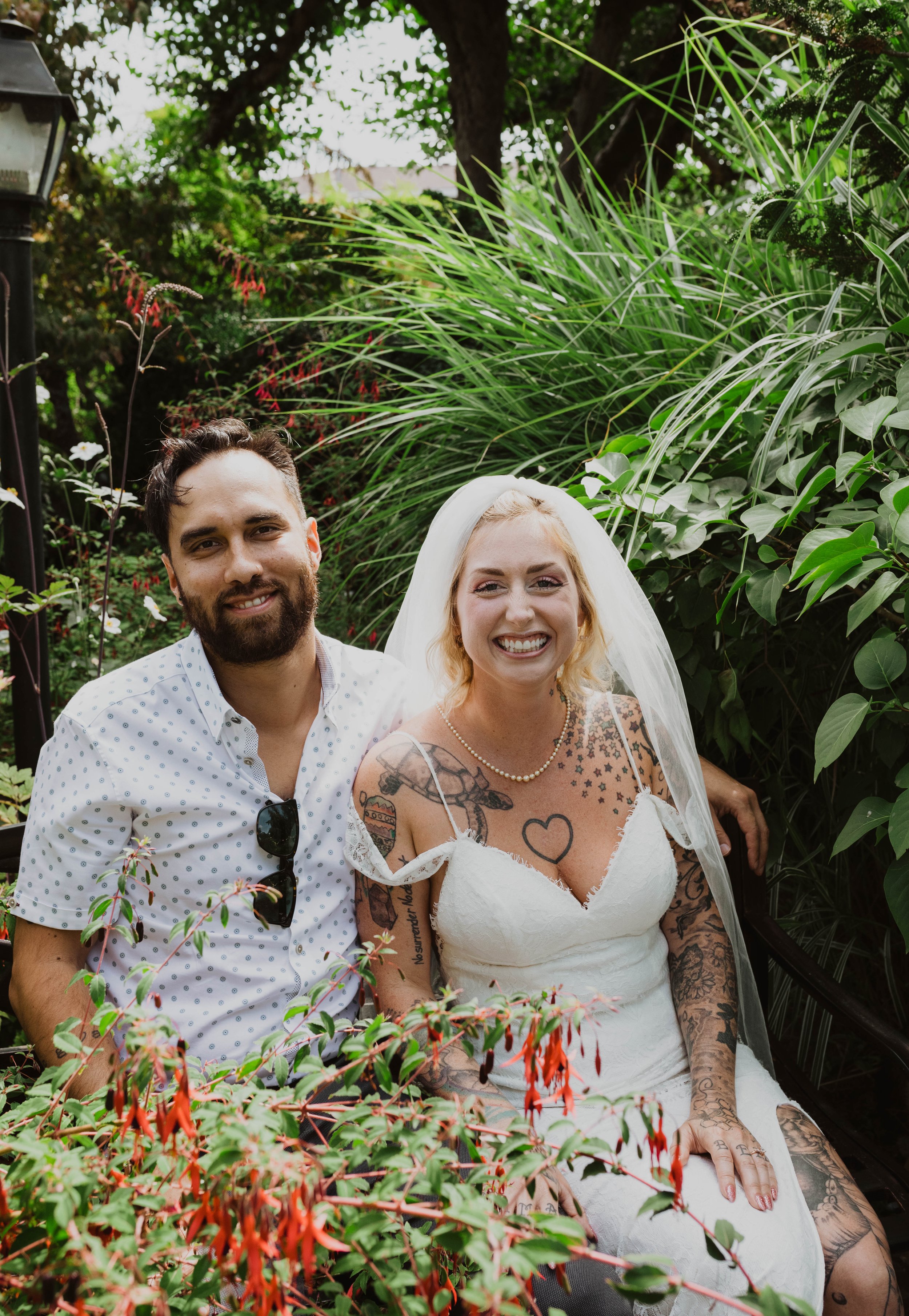 A smiling couple, a man with dark hair and beard in a white polka dot shirt, and a woman with blonde hair in a wedding dress with tattoos, sitting together on a bench amidst lush greenery. Seattle, WA wedding photography.