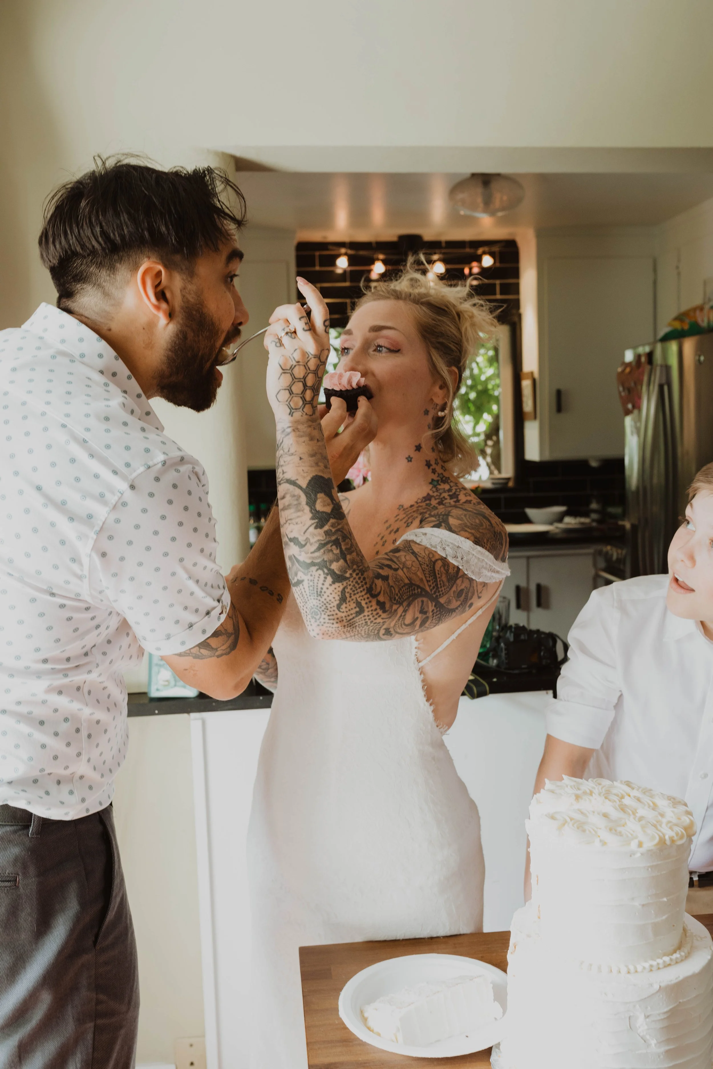 Woman in white dress with tattoos being fed cake by man, in a kitchen with a boy and wedding cake on table. Seattle, WA wedding photography.