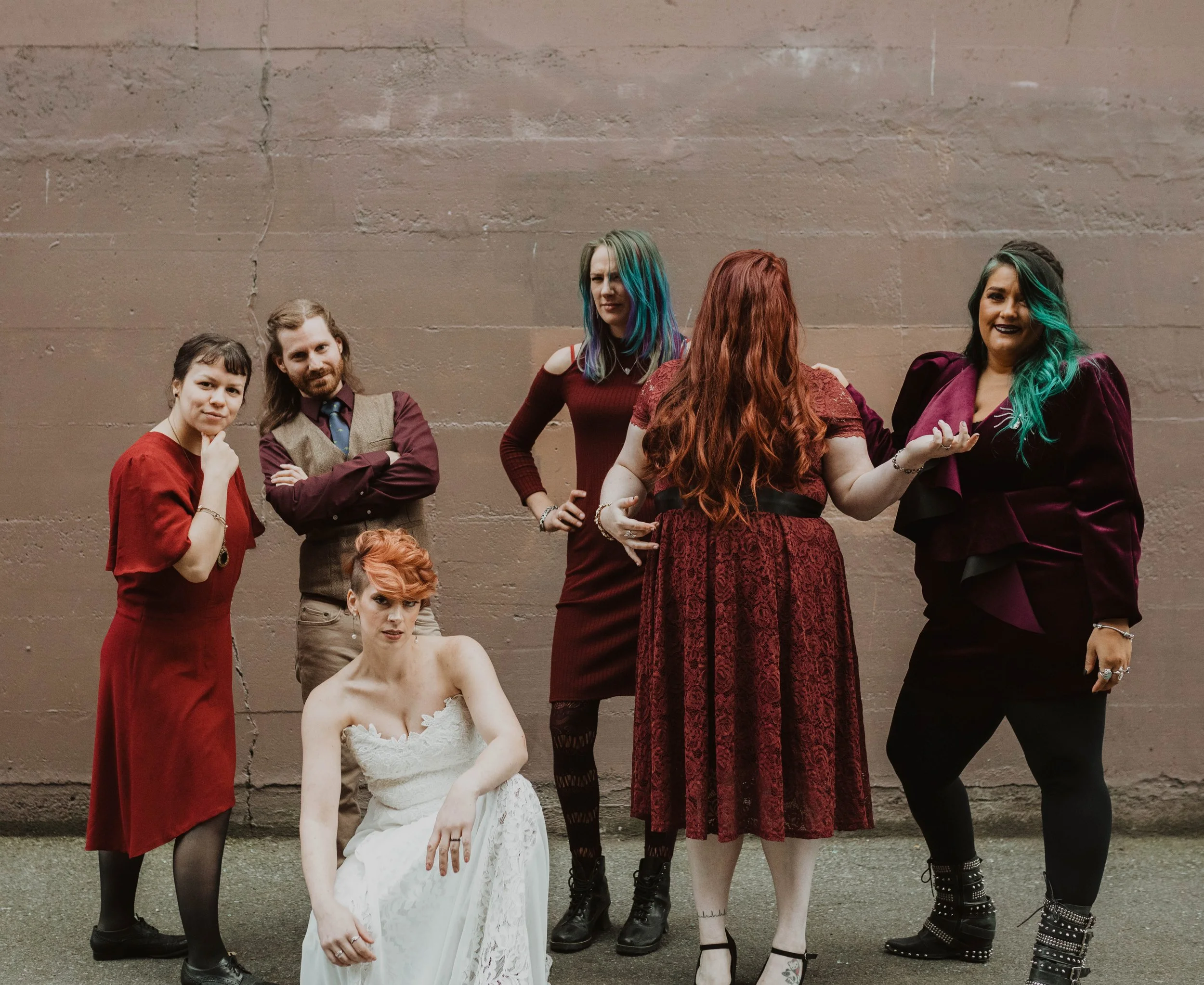 A group of seven women and one man posing against a plain wall, with one woman sitting on the ground wearing a white dress, and others standing around her in various colorful outfits. Pioneer Square, Seattle, WA wedding photography.