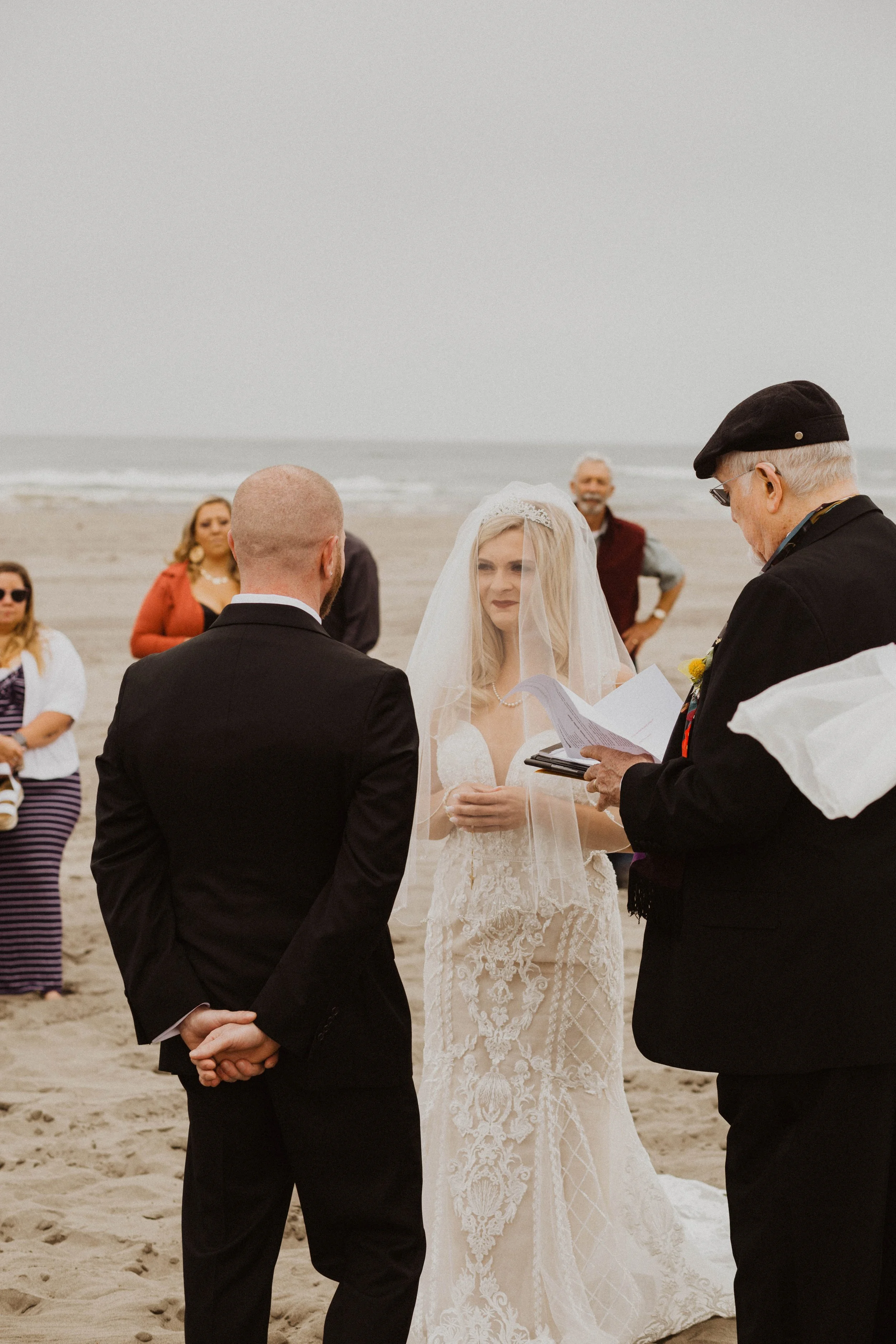 A beach wedding ceremony with a bride in a lace wedding dress and veil, a groom in a black suit, and a officiant reading from papers, surrounded by a few guests during overcast weather. Long Beach, WA wedding photography.