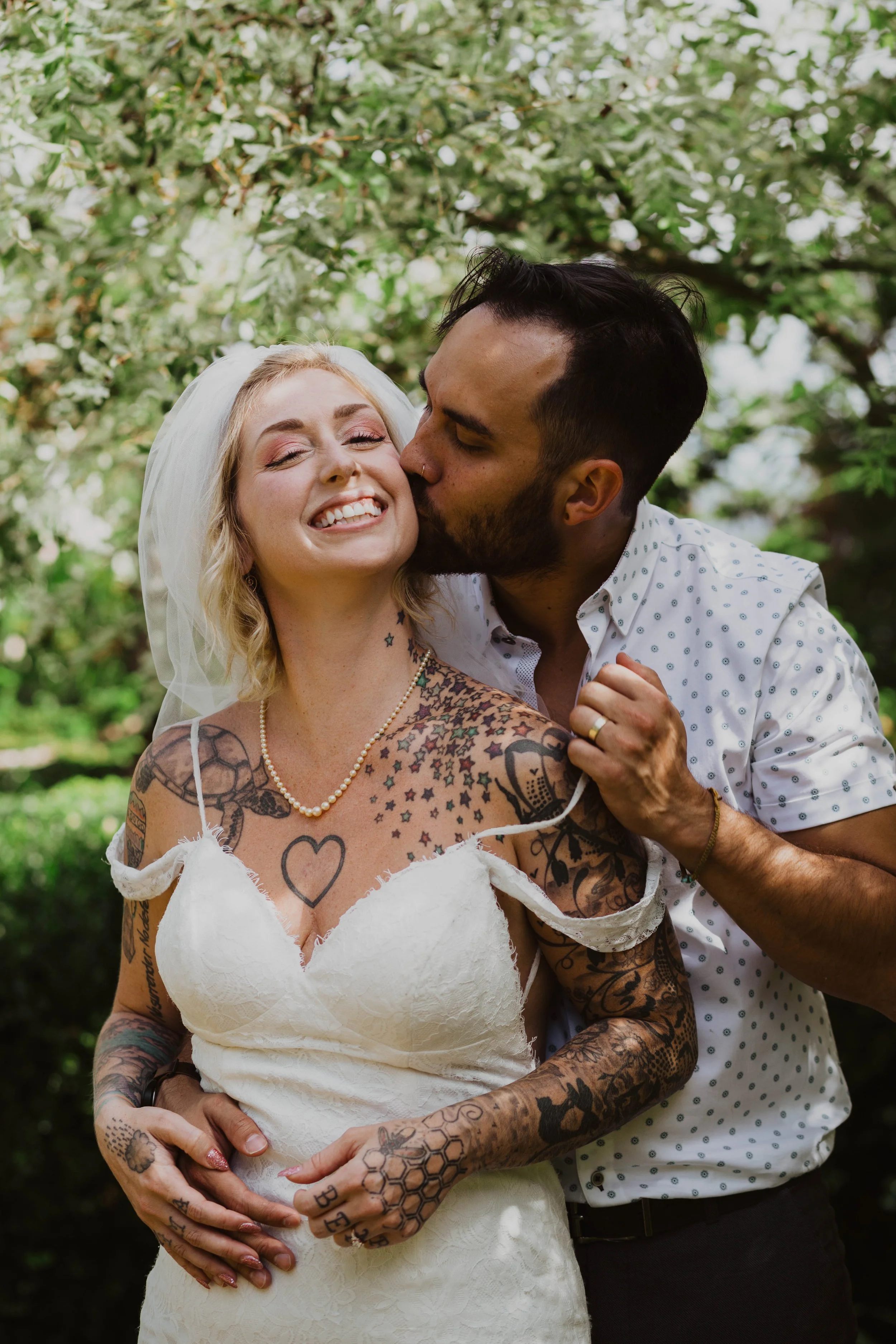 A newlywed couple, the woman with tattoos and a white dress, the man in a white shirt with small blue polka dots, sharing a kiss in a garden with blooming trees. Seattle, WA wedding photography.
