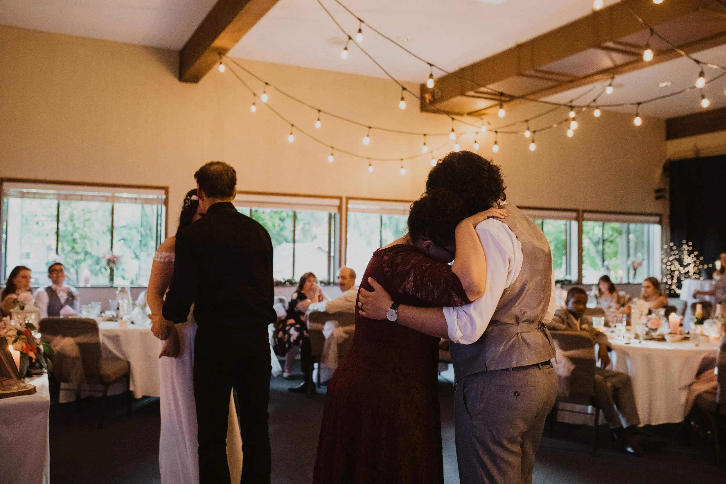 People dancing and hugging at a wedding reception in a decorated hall with string lights and windows showing greenery outside. Seattle, WA wedding photography.