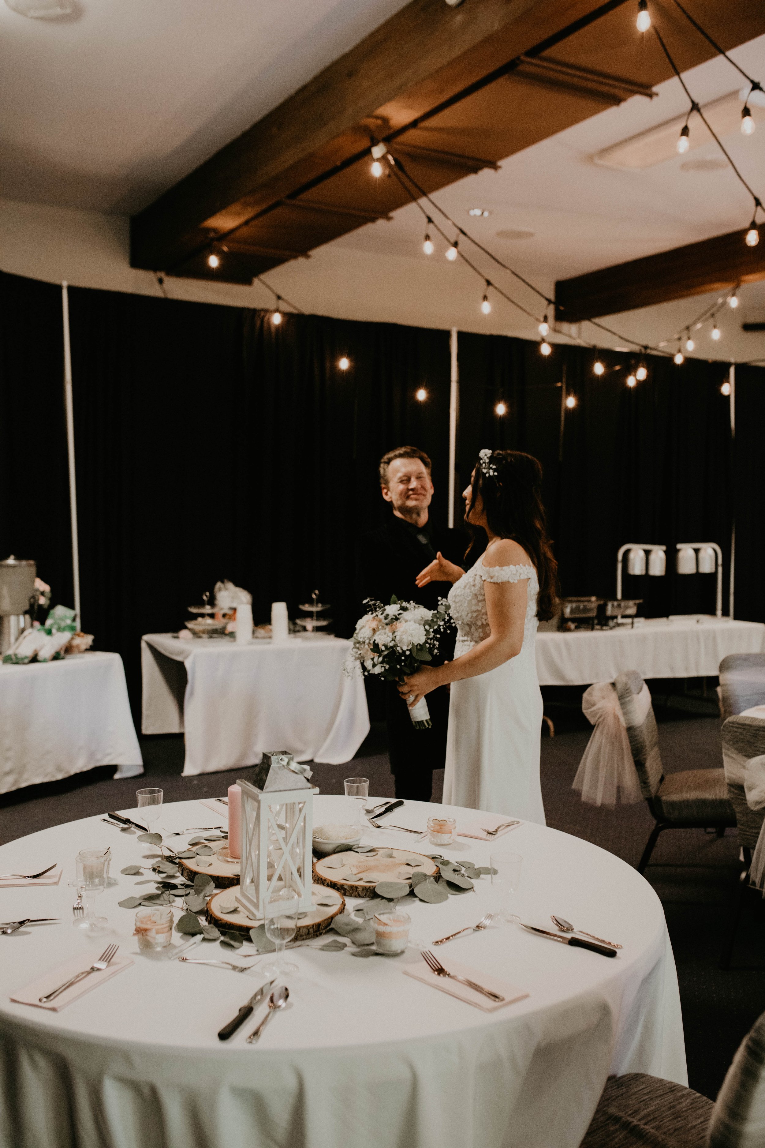 A bride and an older man, possibly her father, sharing a dance at a wedding reception in a decorated banquet hall with string lights and a table set for dinner. Seattle, WA wedding photography. 