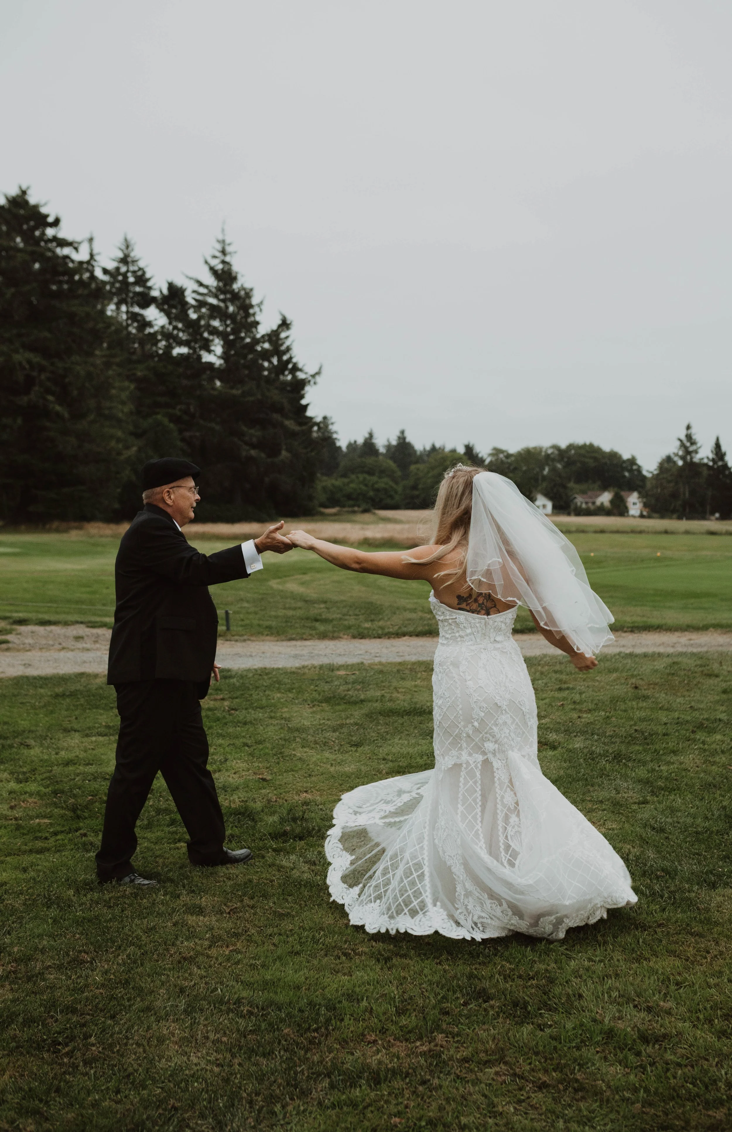 A bride in a white lace wedding dress and veil is holding hands with an elderly man dressed in a black suit and hat outside on a grassy field, with trees and a cloudy sky in the background. Long Beach, WA wedding photography.