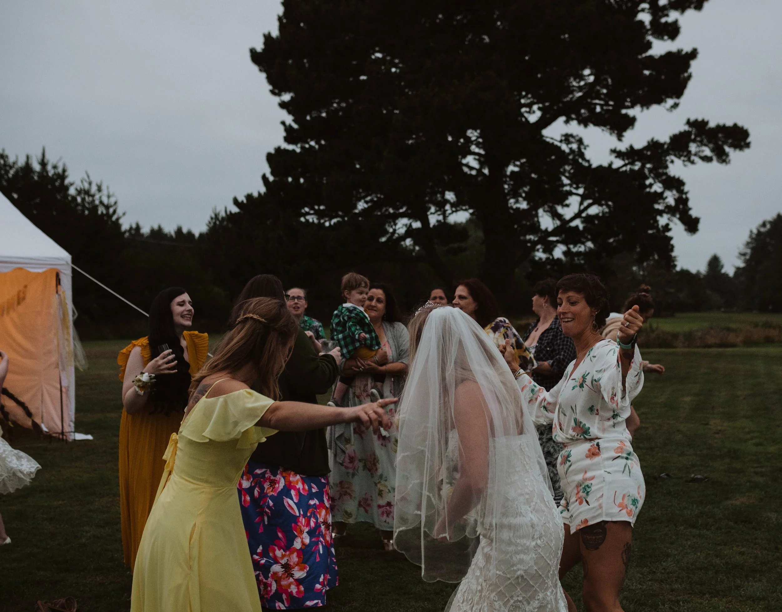 Group of women and a child dancing and celebrating outdoors at a wedding reception, with a bride in a white dress and veil, in a grassy area with trees at dusk. Long Beach, WA wedding photography.