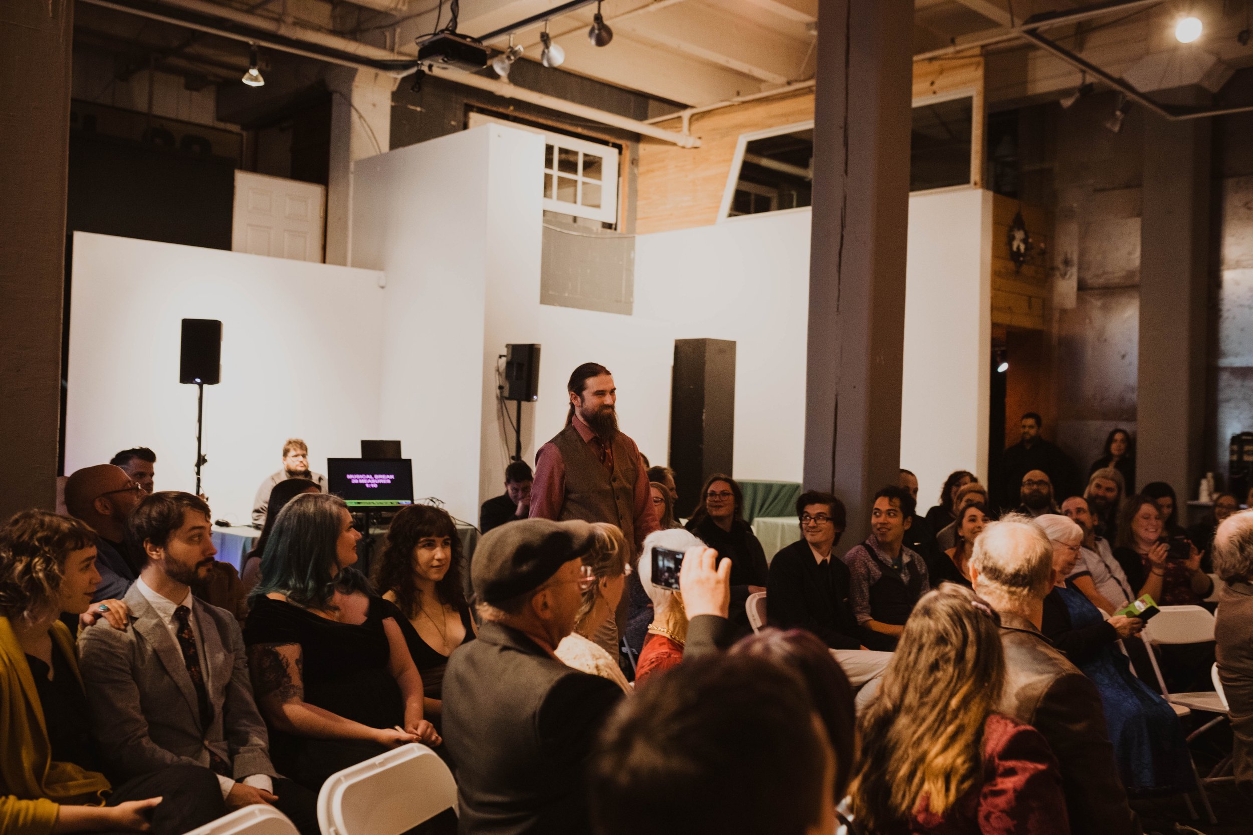 Audience attending a presentation or event in a modern venue with white walls and industrial decor, with a man standing and speaking among seated attendees. Pioneer Square, Seattle, WA wedding photography.