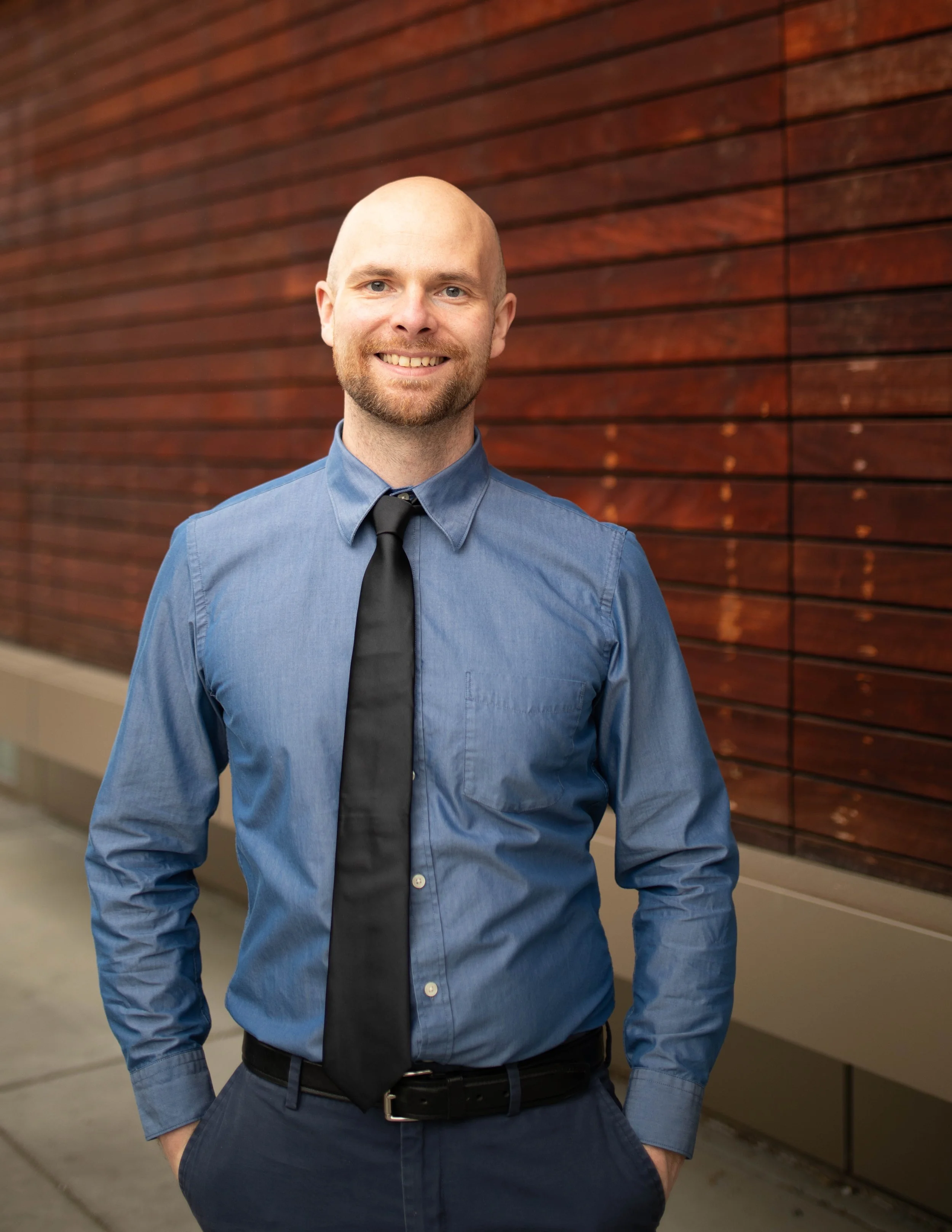 A man standing outside in front of a wooden panel wall, wearing a blue dress shirt and a black tie, smiling with hands in pockets. Seattle professional head shot photography