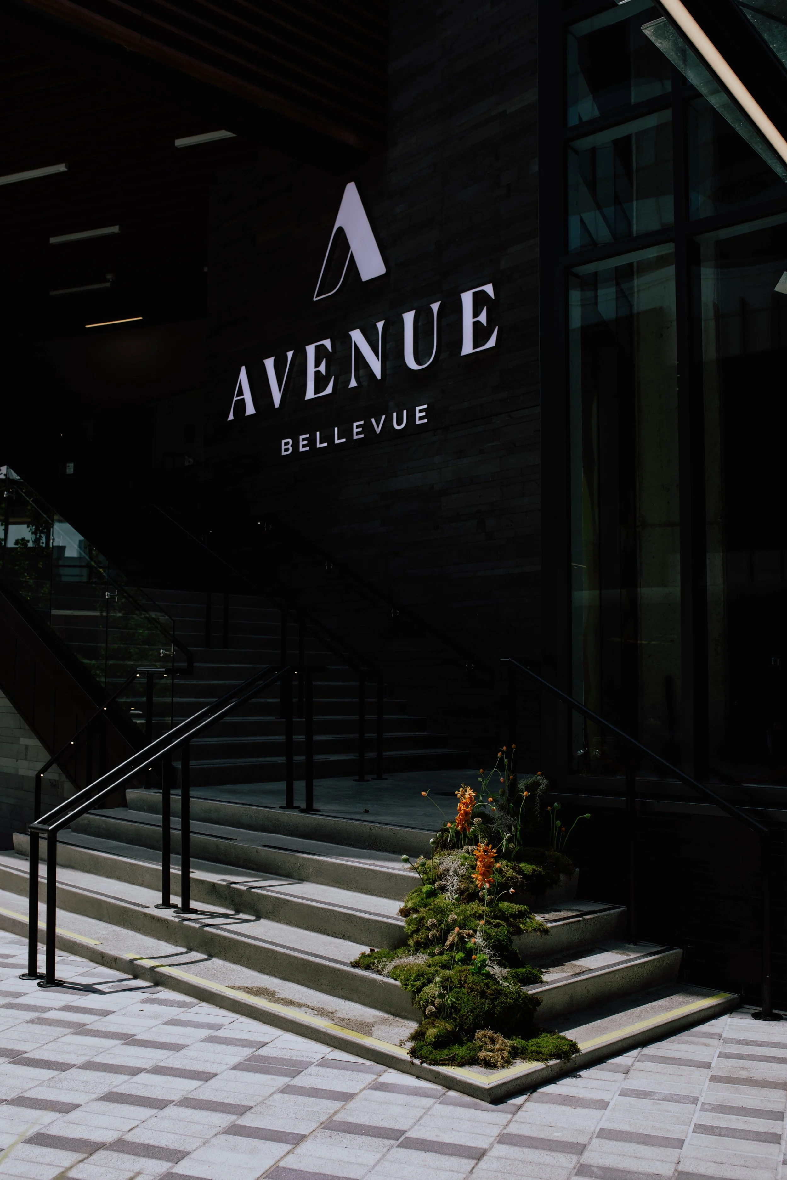 Exterior view of Avenue Bellevue entrance with stairs, black handrails, and a flower arrangement on the step, illuminated sign displaying 'Avenue Bellevue' against a dark wall. Seattle event photography