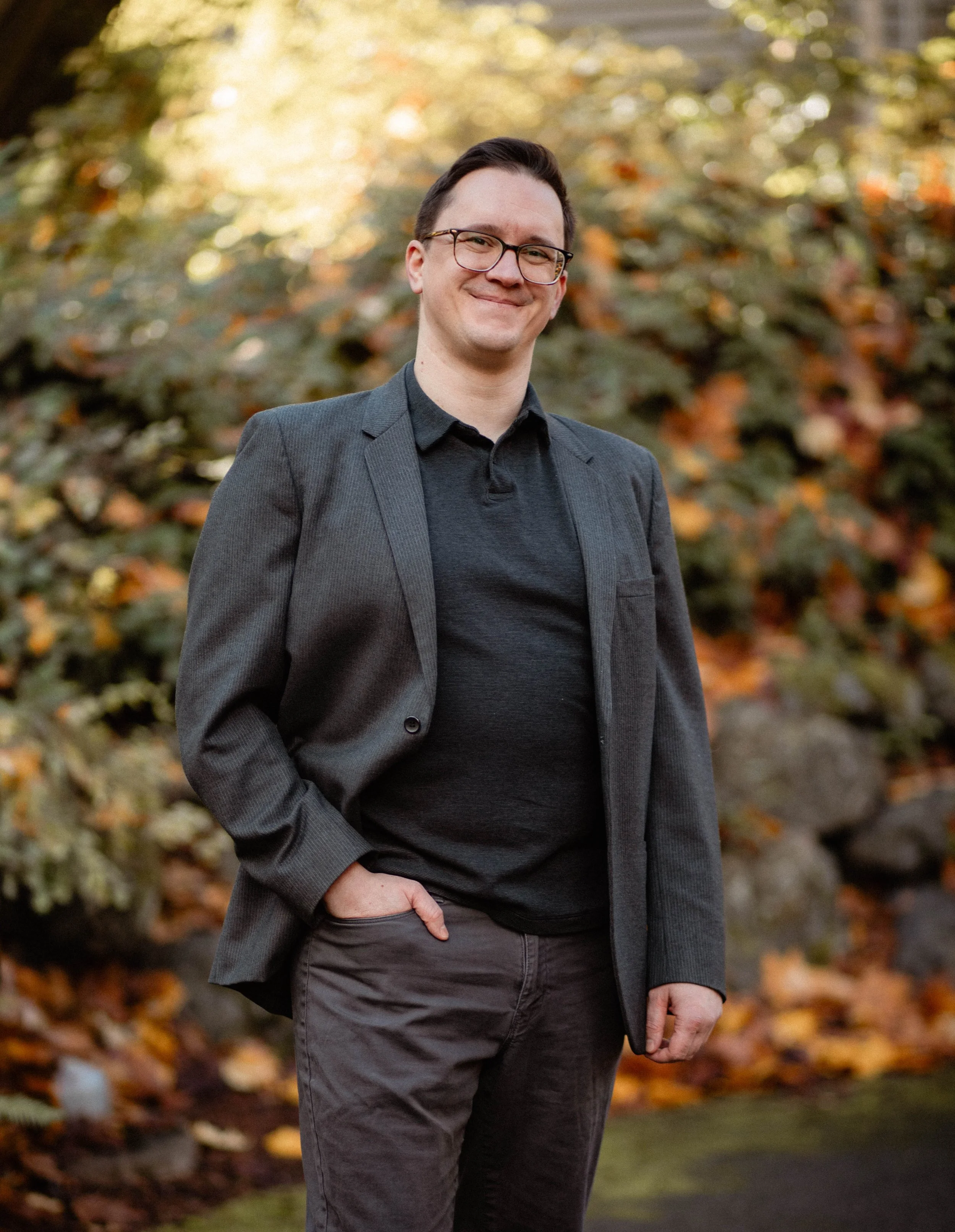 A man wearing glasses, a gray blazer, and dark clothing standing outdoors in front of autumn foliage. Seattle professional head shot photography