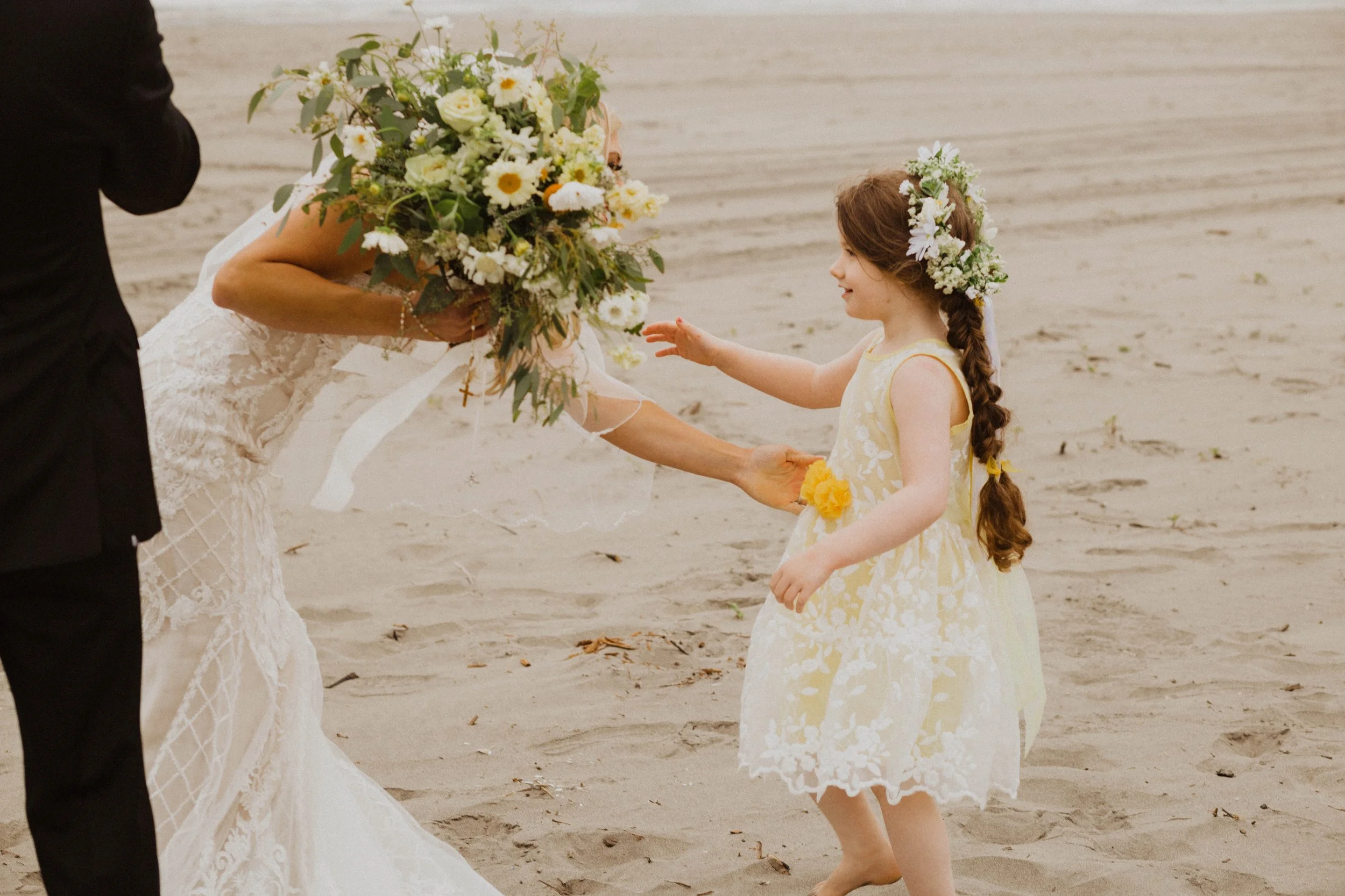 A little girl with a braid and floral headpiece reaching for a bride's bouquet on a beach. Long Beach, WA wedding photography.