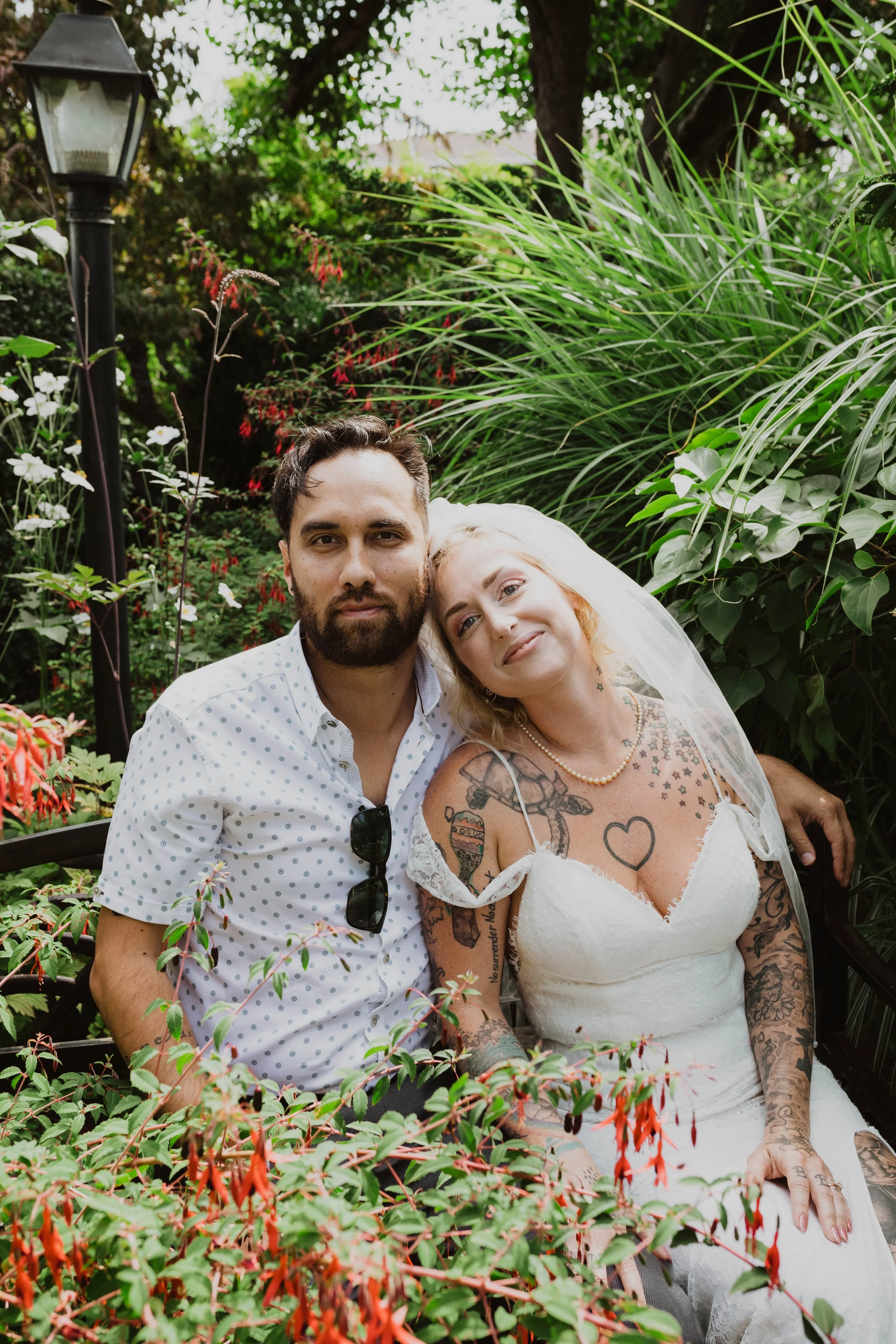 A couple sitting on a bench surrounded by lush green plants and flowers, with the woman wearing a wedding dress and the man in a white shirt with sunglasses hanging from the collar. Seattle, WA wedding photography.