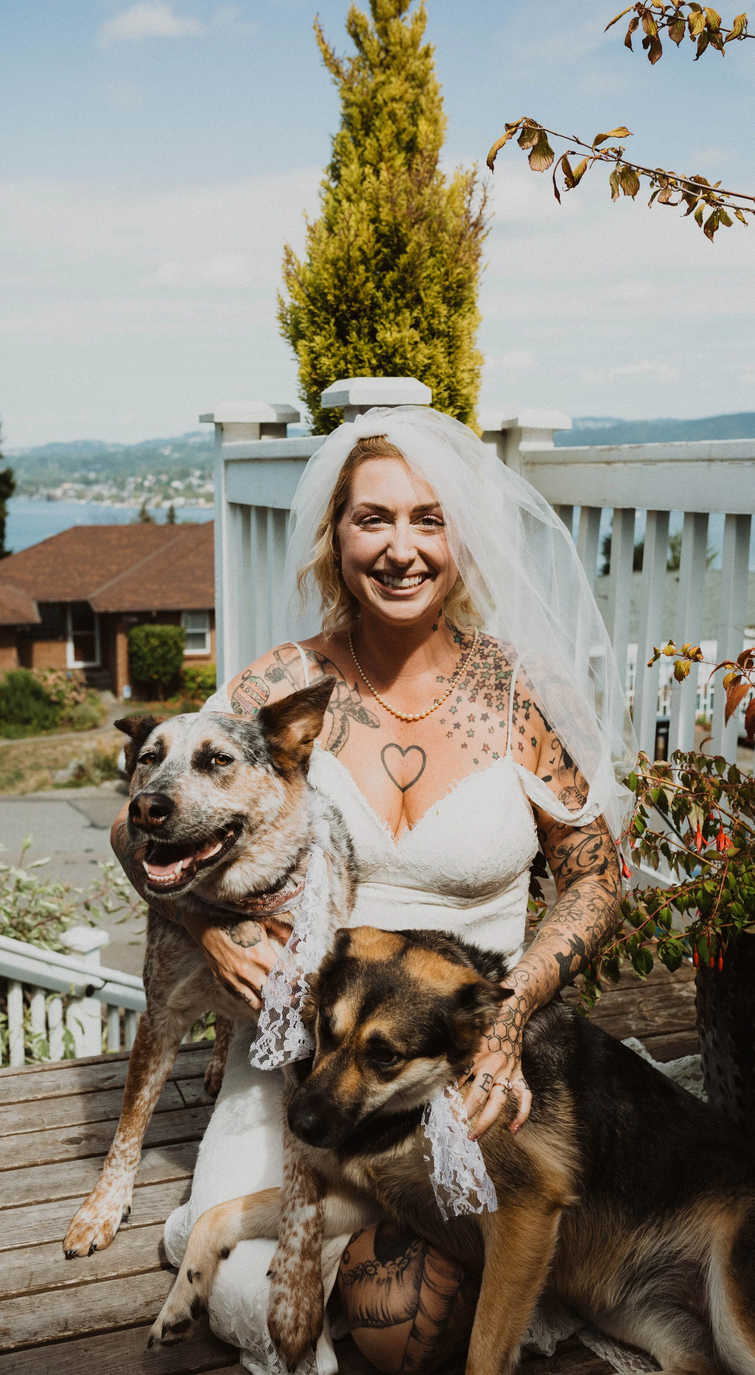 A smiling woman dressed as a bride with tattoos, wearing a wedding dress and veil, sitting outdoors with two dogs, one white and brindle, one black and tan, on a wooden deck with a railing, in a residential area near a body of water and greenery.