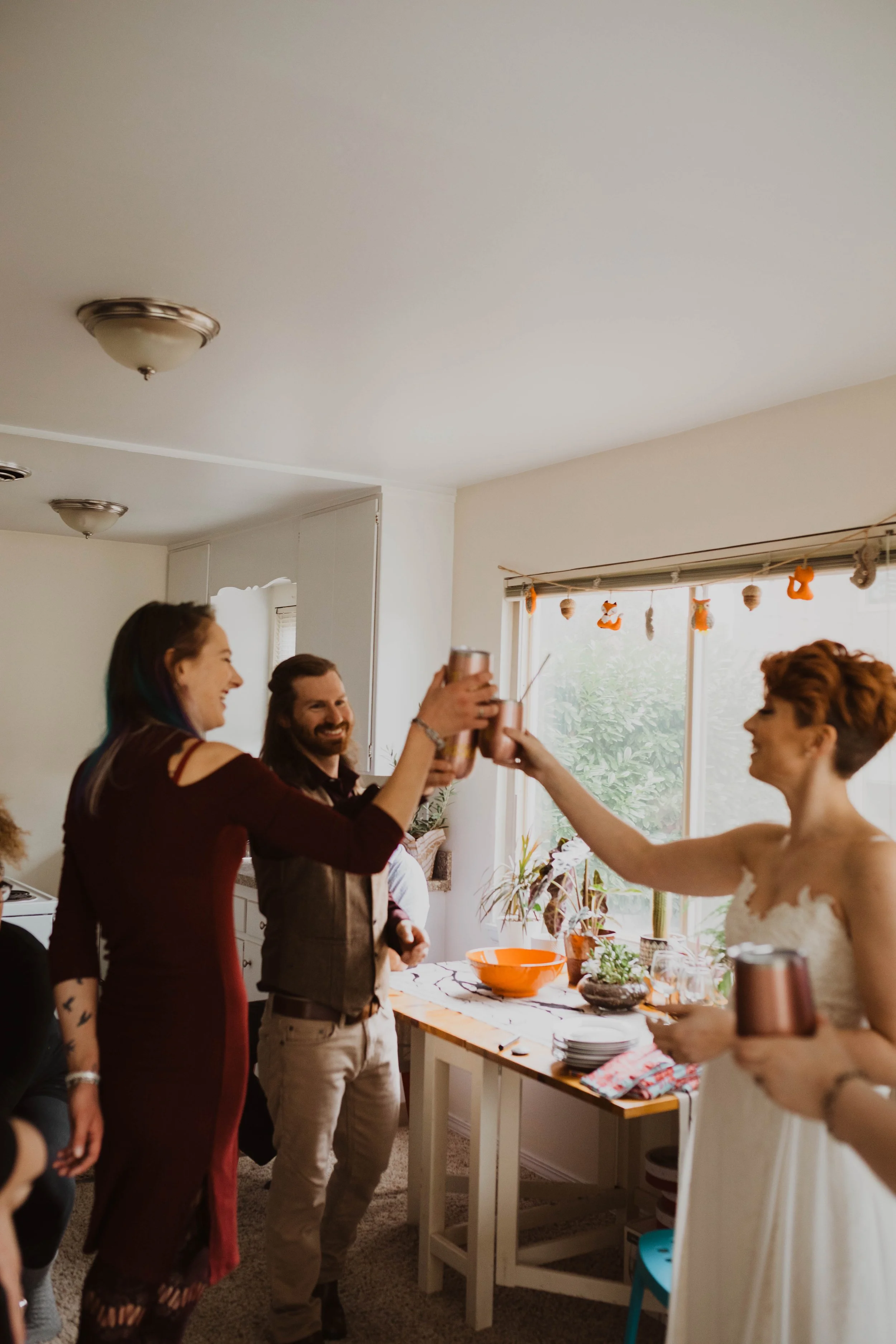 People celebrating, clinking drinks at a gathering inside a house, decorated with small hanging decorations near the window. Pioneer Square, Seattle, WA wedding photography.