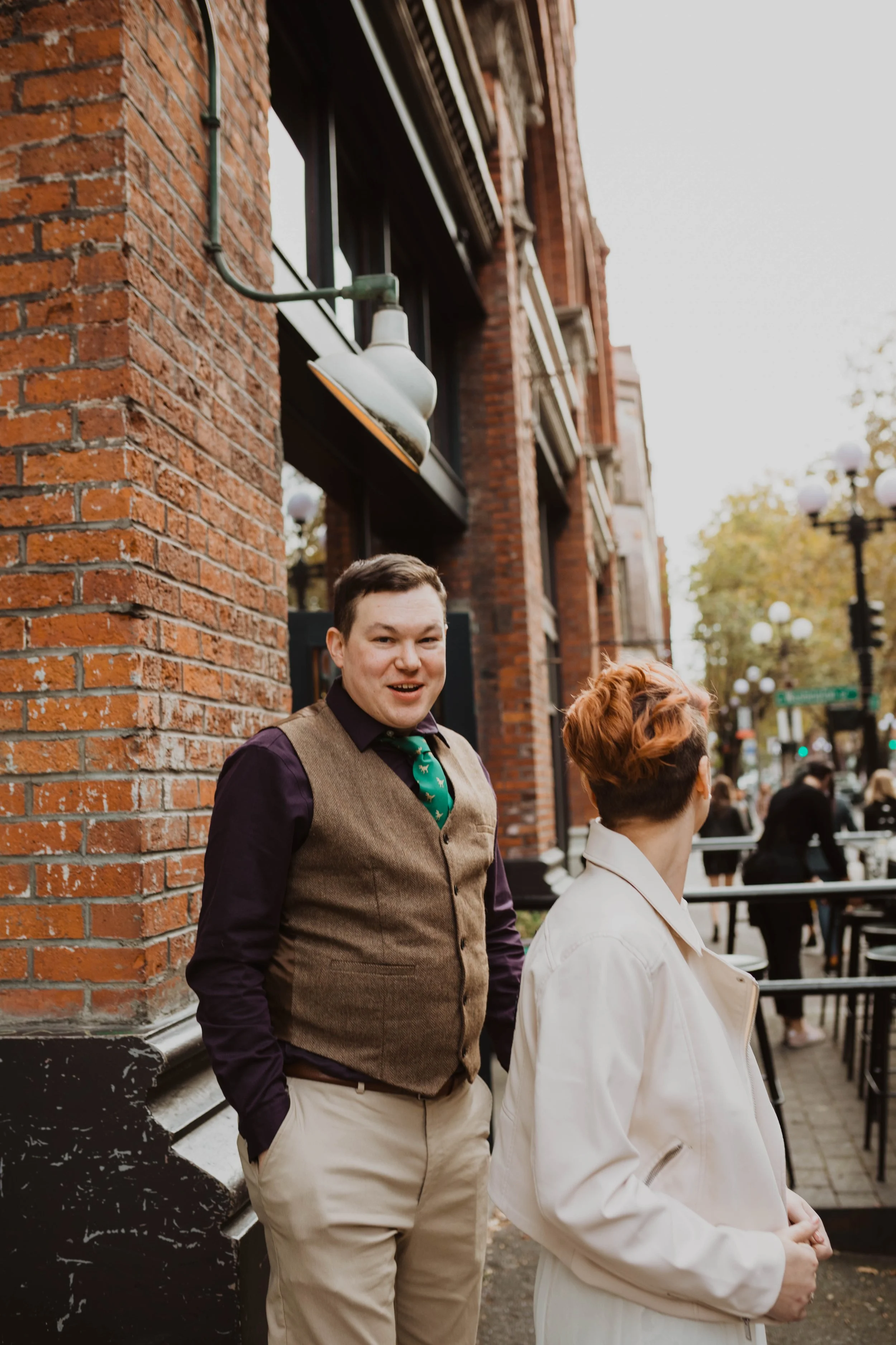 Two people standing outside near a brick building with outdoor seating, street lamps, and trees in the background. Pioneer Square, Seattle, WA wedding photography.
