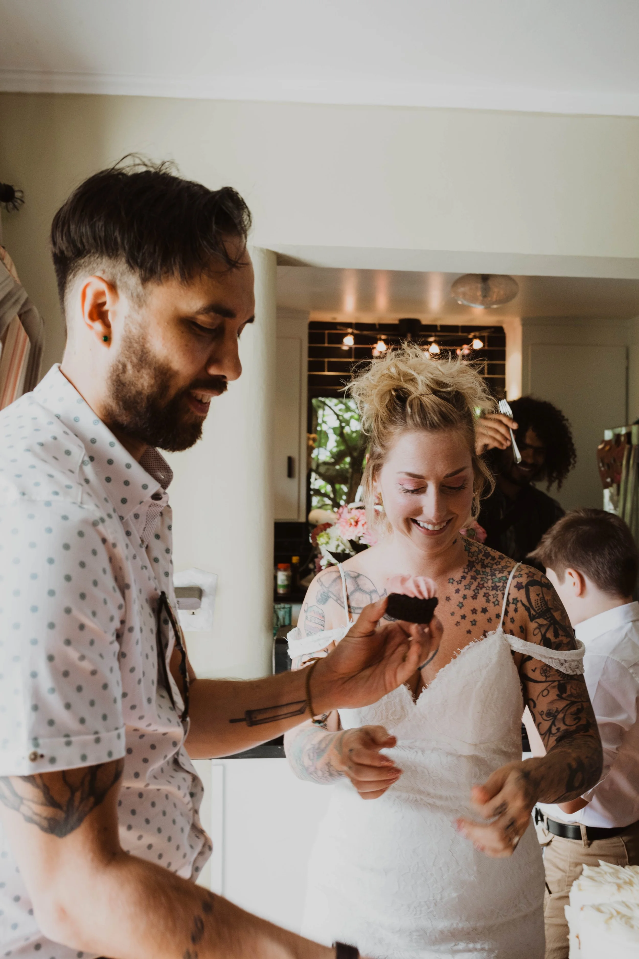 A man and woman sharing a moment as the man offers her a piece of chocolate cake, with other party guests in the background. Seattle, WA wedding photography.