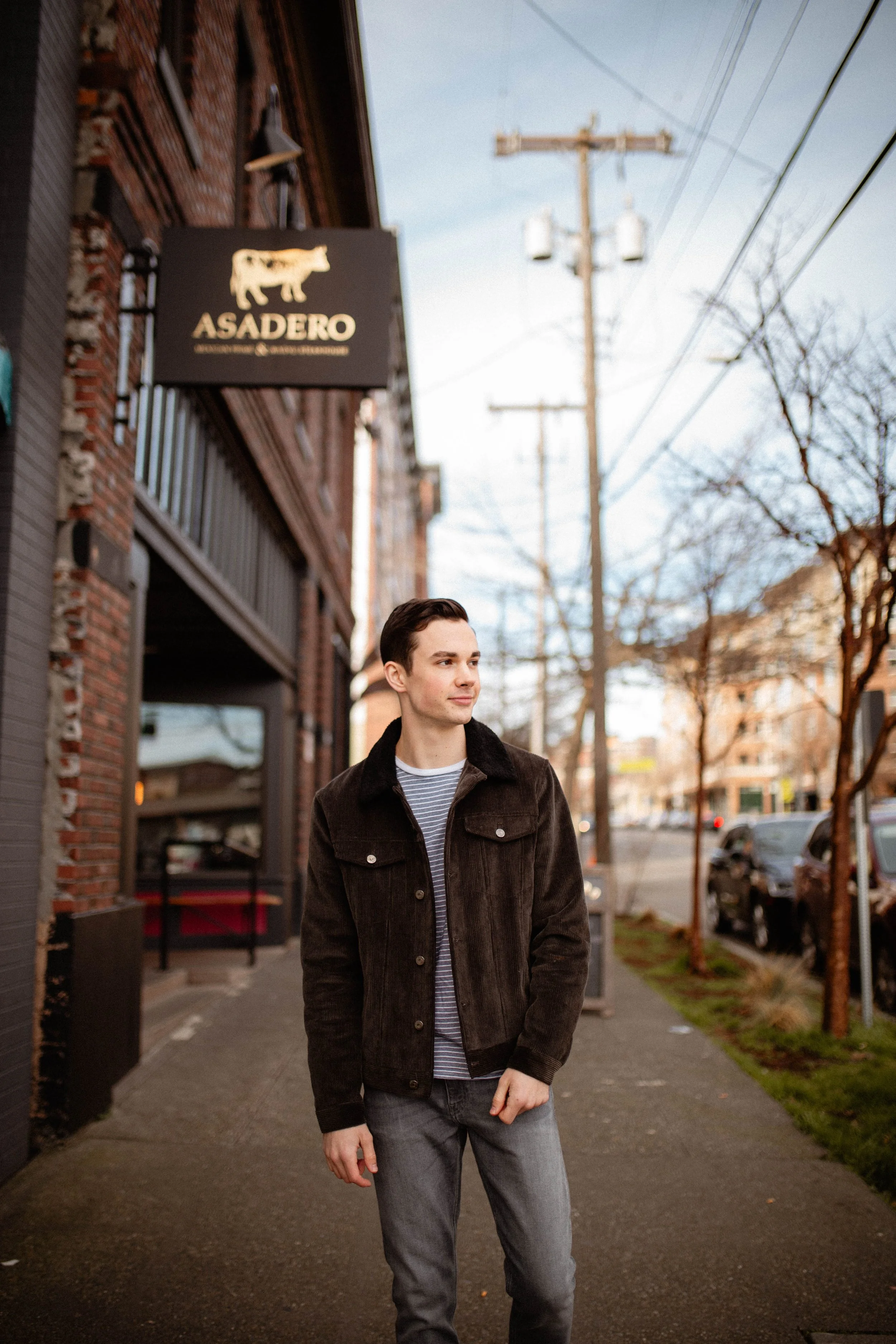 Young man walking on city sidewalk on a clear day, with brick building and restaurant sign reading 'Asadero' in background. Seattle professional head shot photography