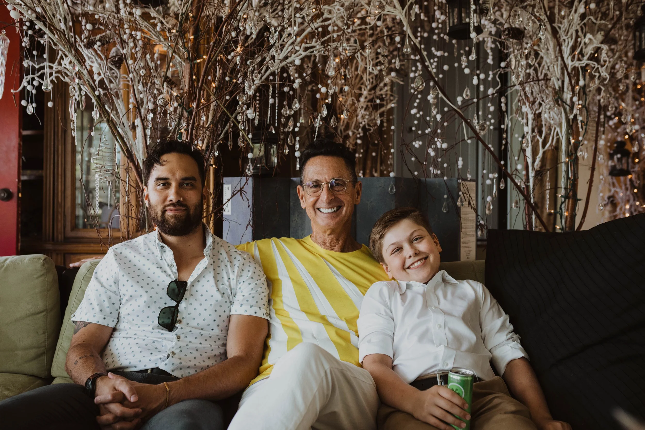 Three males sitting on a couch in a decorated indoor space, smiling at the camera.  The man on the left has a beard, dark hair, and sunglasses hanging on his shirt. The man in the middle is wearing glasses and a yellow and white striped shirt. The yo