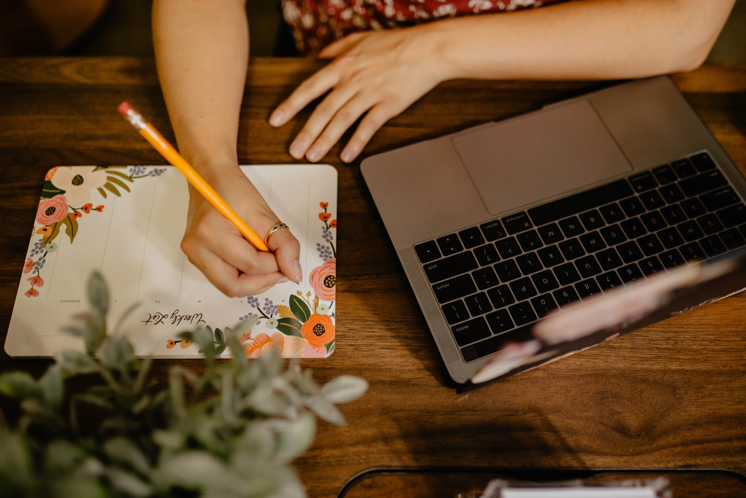 A person writes on a floral decorated weekly planner with a yellow pencil, placed on a wooden table next to a gray laptop. Seattle professional head shot photography