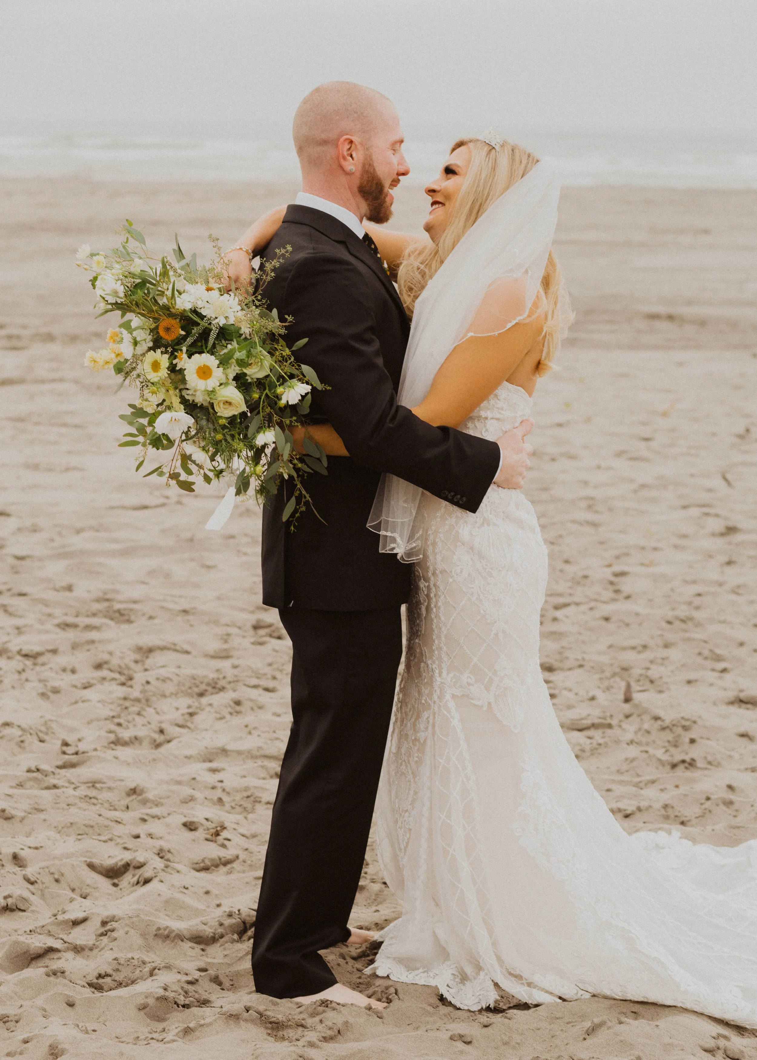 A bride and groom embrace on a sandy beach, facing each other and smiling with the ocean in the background. The groom holds a large bouquet of flowers, and the bride is wearing a white wedding dress with a veil. Long Beach, WA wedding photography.