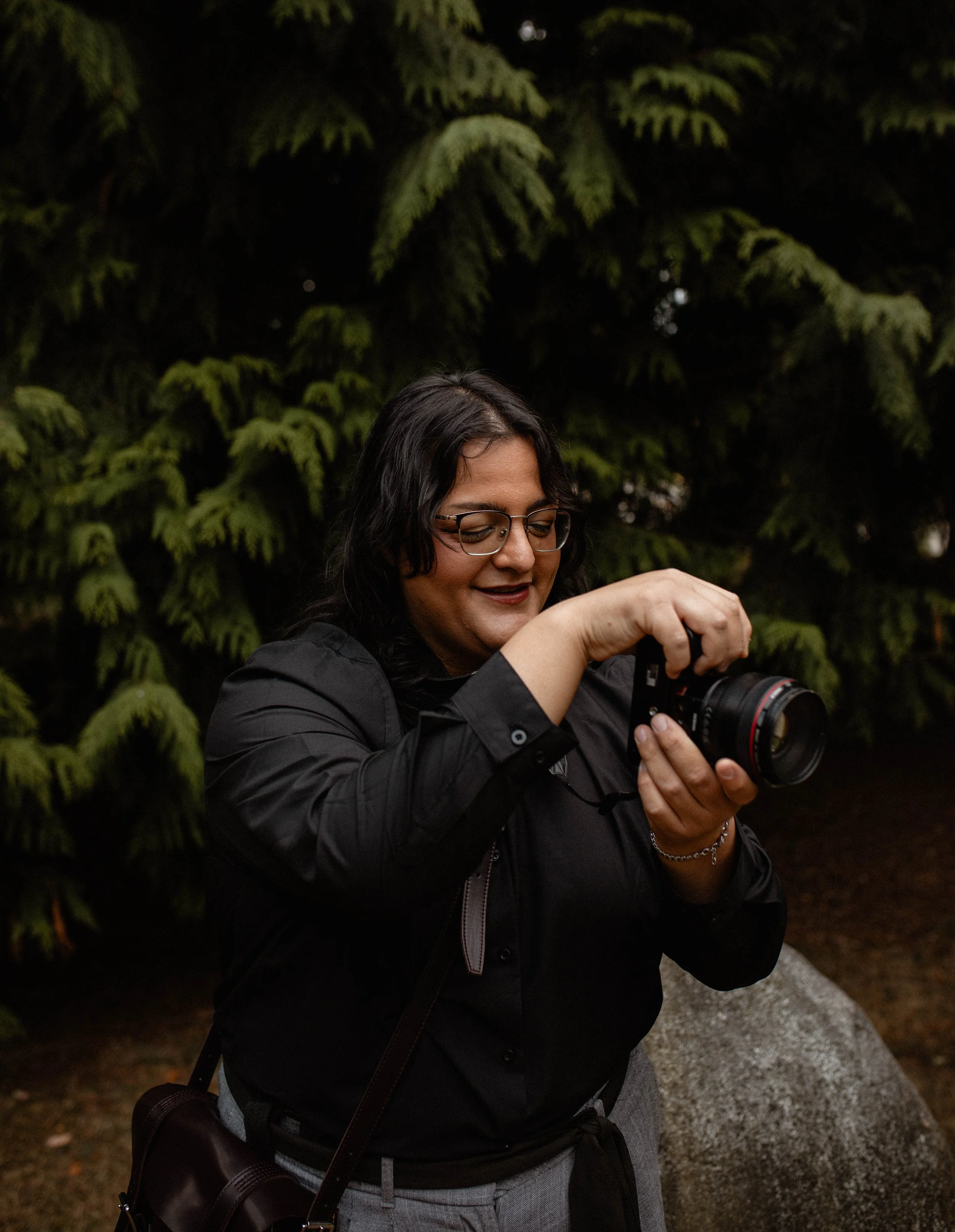 A woman with dark hair, glasses, and a black jacket is holding a camera outdoors near a large rock and evergreen trees. Seattle professional head shot photography
