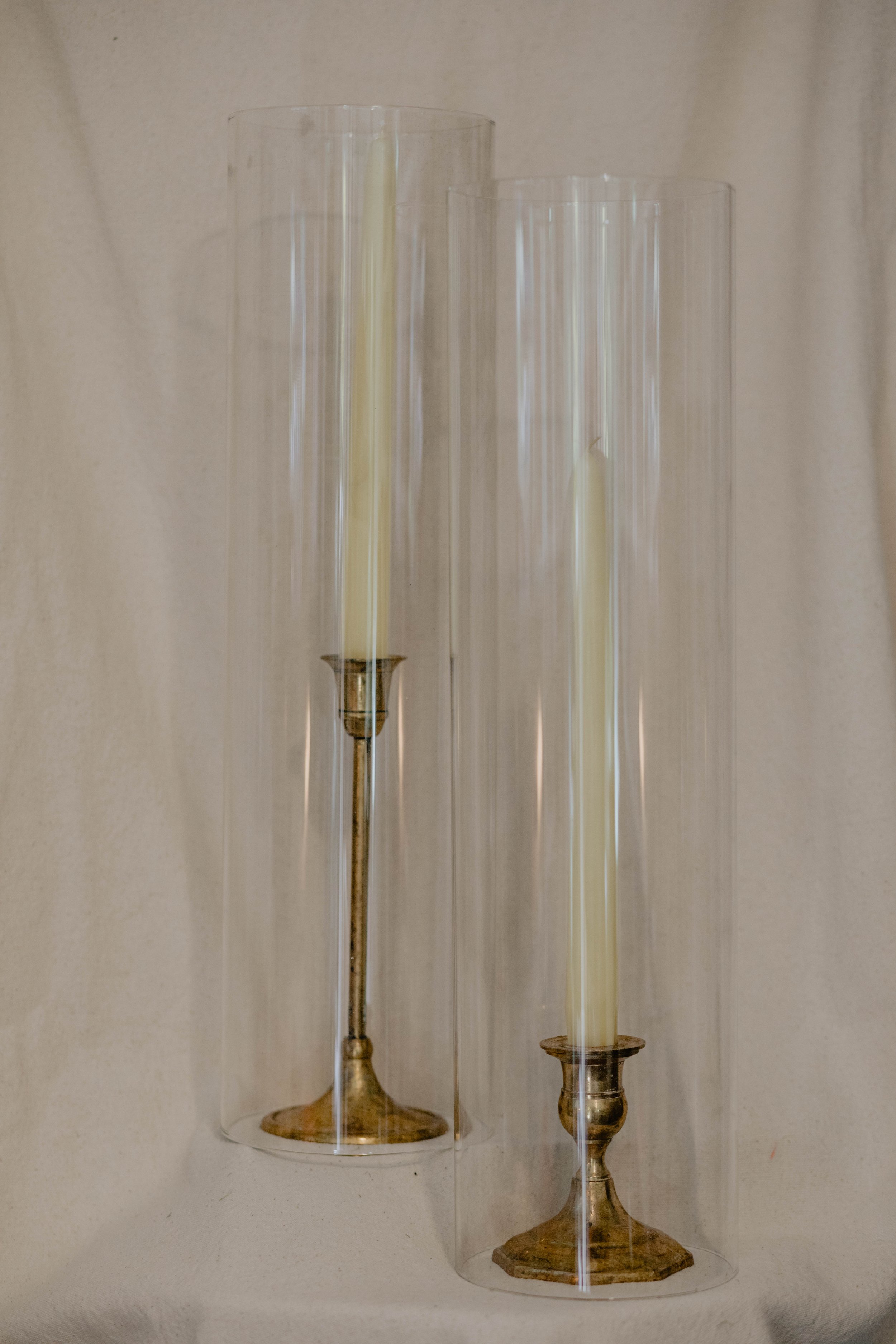 Two tall candlesticks with long white candles, encased in clear cylindrical glass covers, placed on a white surface against a light-colored wall.  Seattle product photography