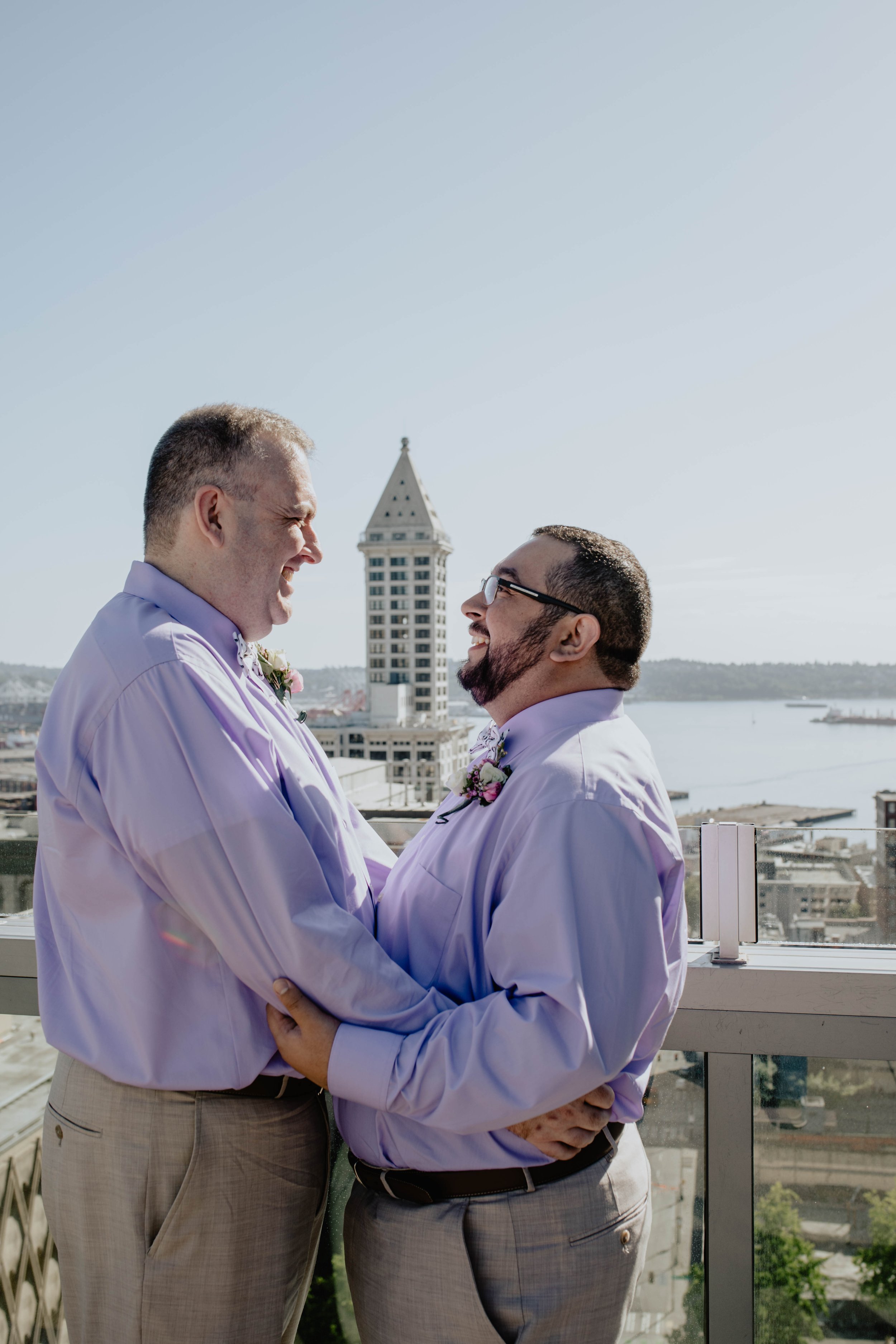 Two men holding each other and smiling on a balcony with a city skyline and waterfront in the background. Seattle Municipal Courthouse wedding photography.