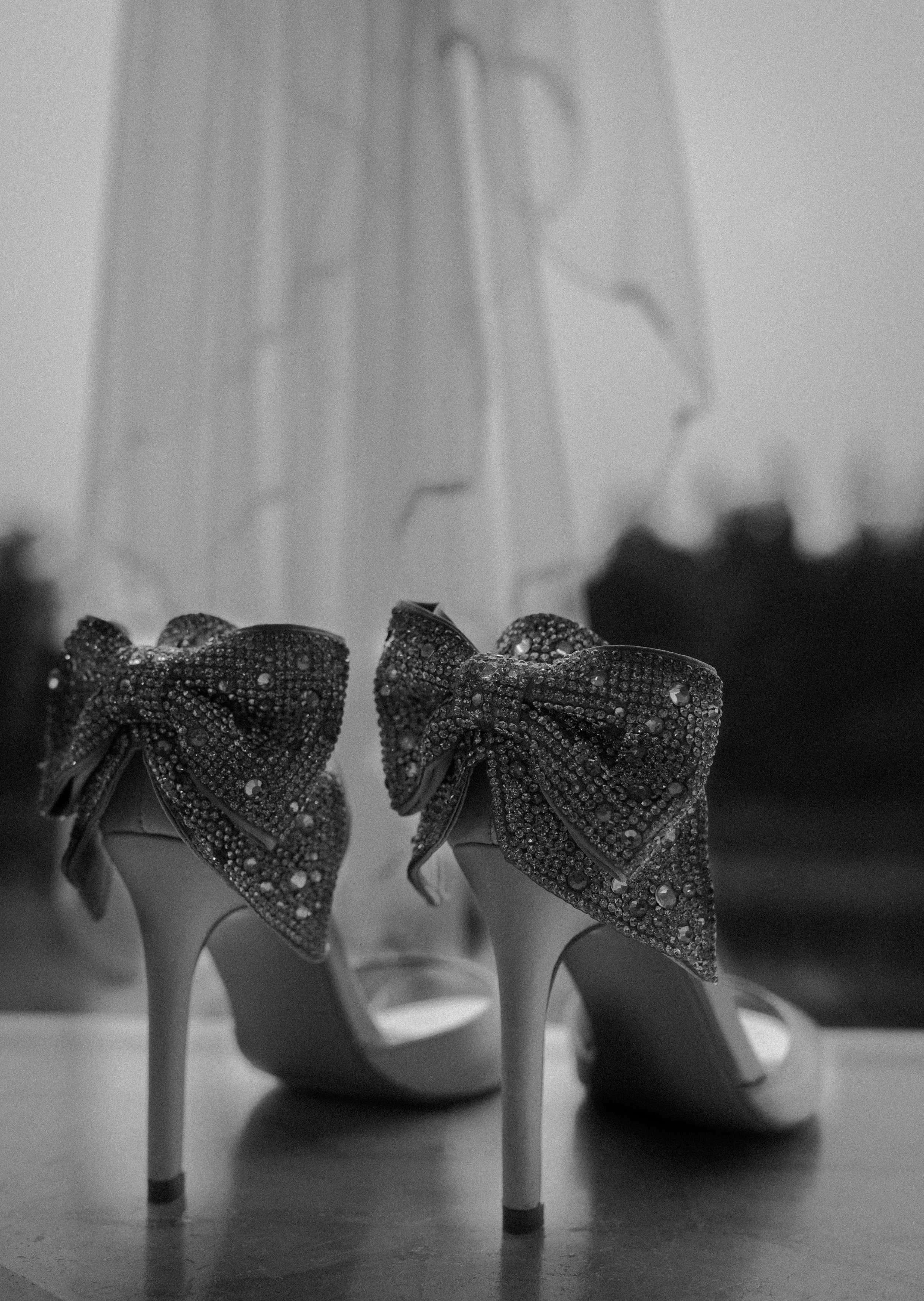 A pair of high-heeled shoes with large, embellished bows on the back, covered in rhinestones, placed on a flat surface. Long Beach, WA wedding photography.