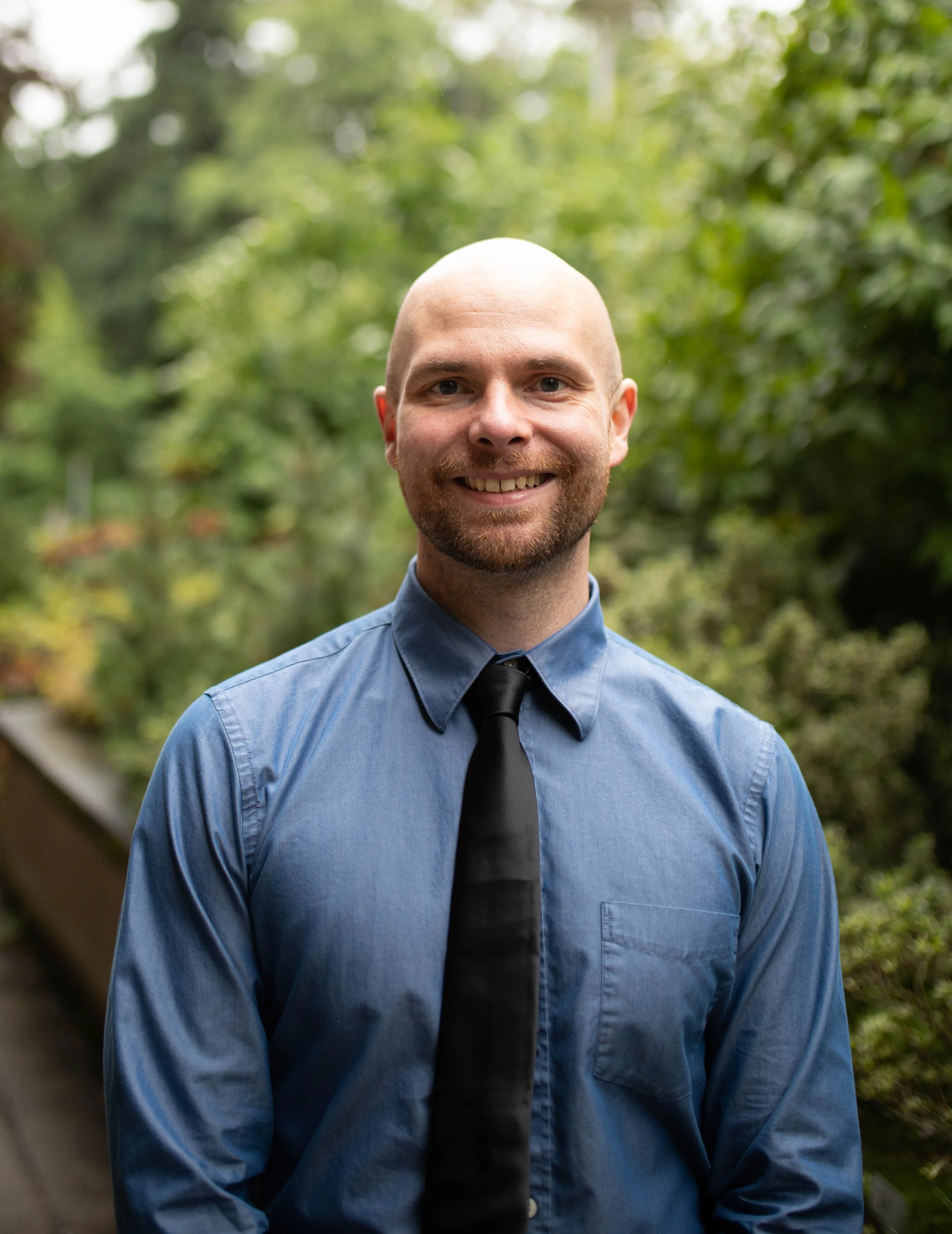 Smiling man with a beard and no hair, wearing a blue dress shirt and black tie, outdoors with green trees in the background. Seattle professional head shot photography