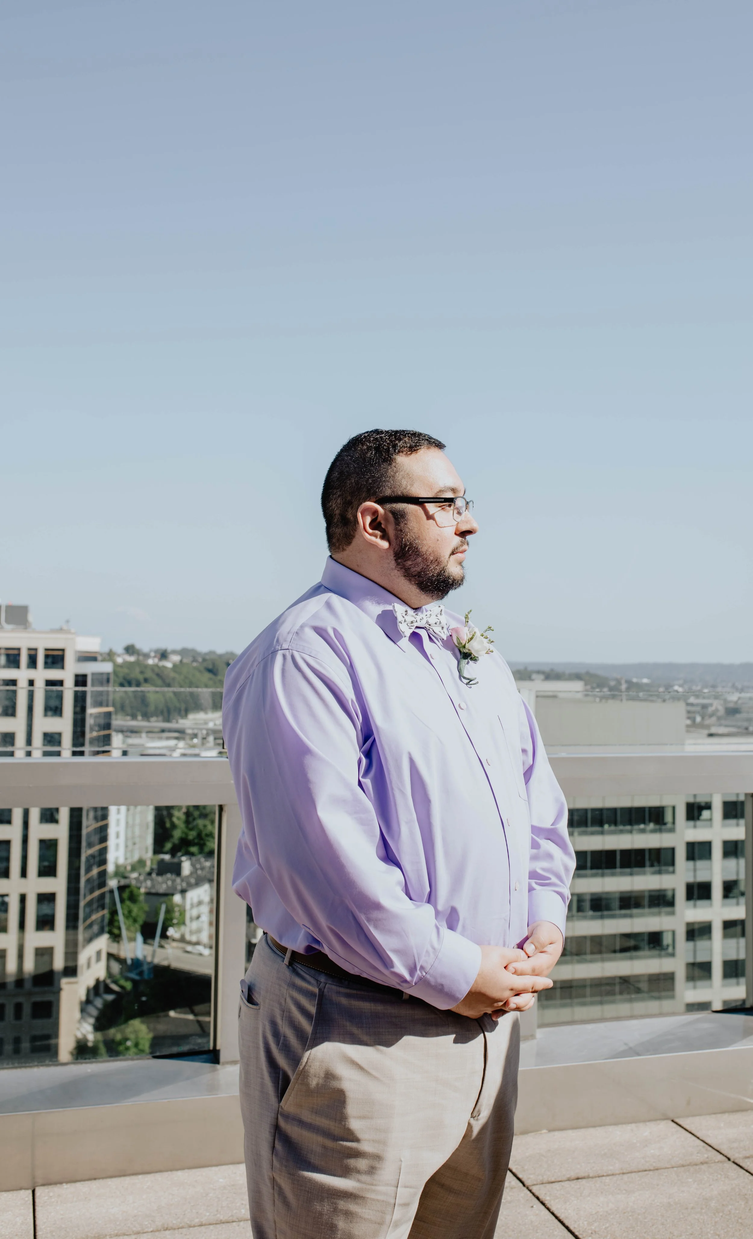 A man with glasses and a beard standing on a rooftop with city buildings in the background, dressed in a light purple shirt and beige pants, with a flower boutonniere. Seattle Municipal Courthouse wedding photography.