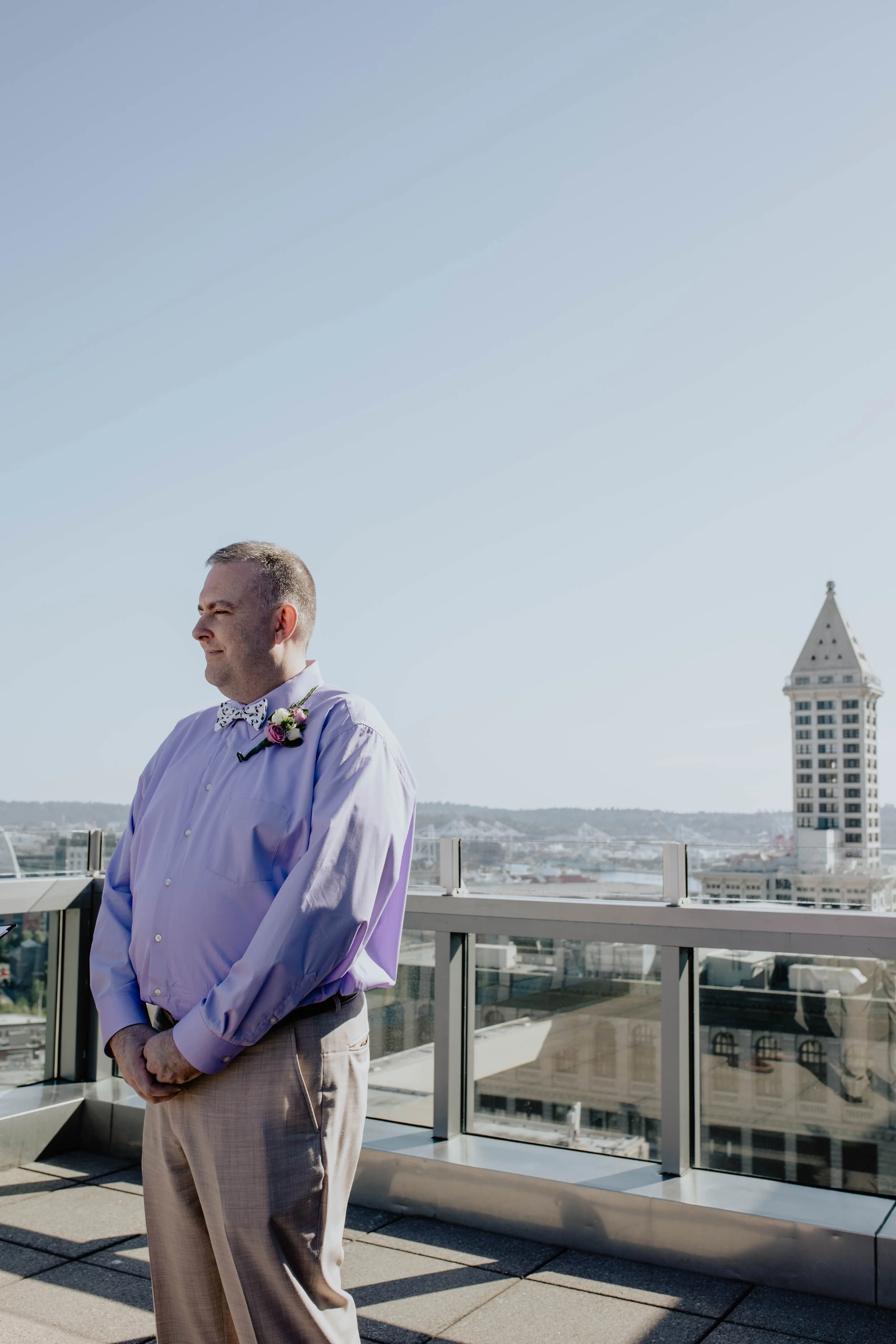 A man dressed in a lavender shirt with a floral bow tie and boutonniere, standing on a rooftop with a city skyline and a tall building in the background, during daytime. Seattle Municipal Courthouse wedding photography.