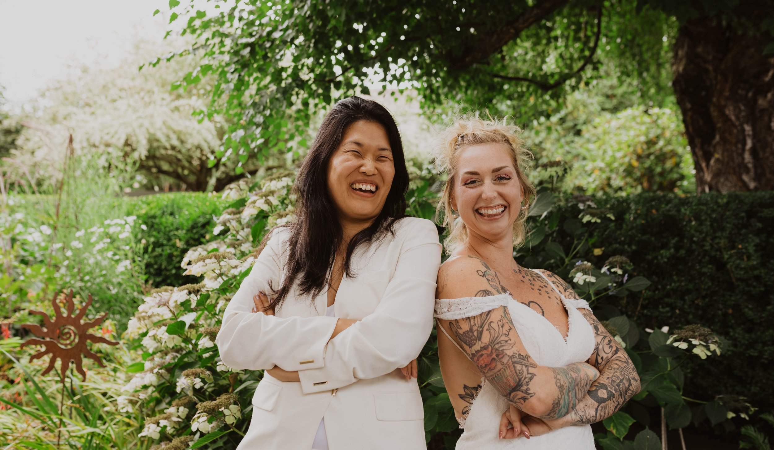 Two women standing outdoors in a garden, smiling and laughing, surrounded by lush greenery and white flowers. Seattle, WA wedding photography.