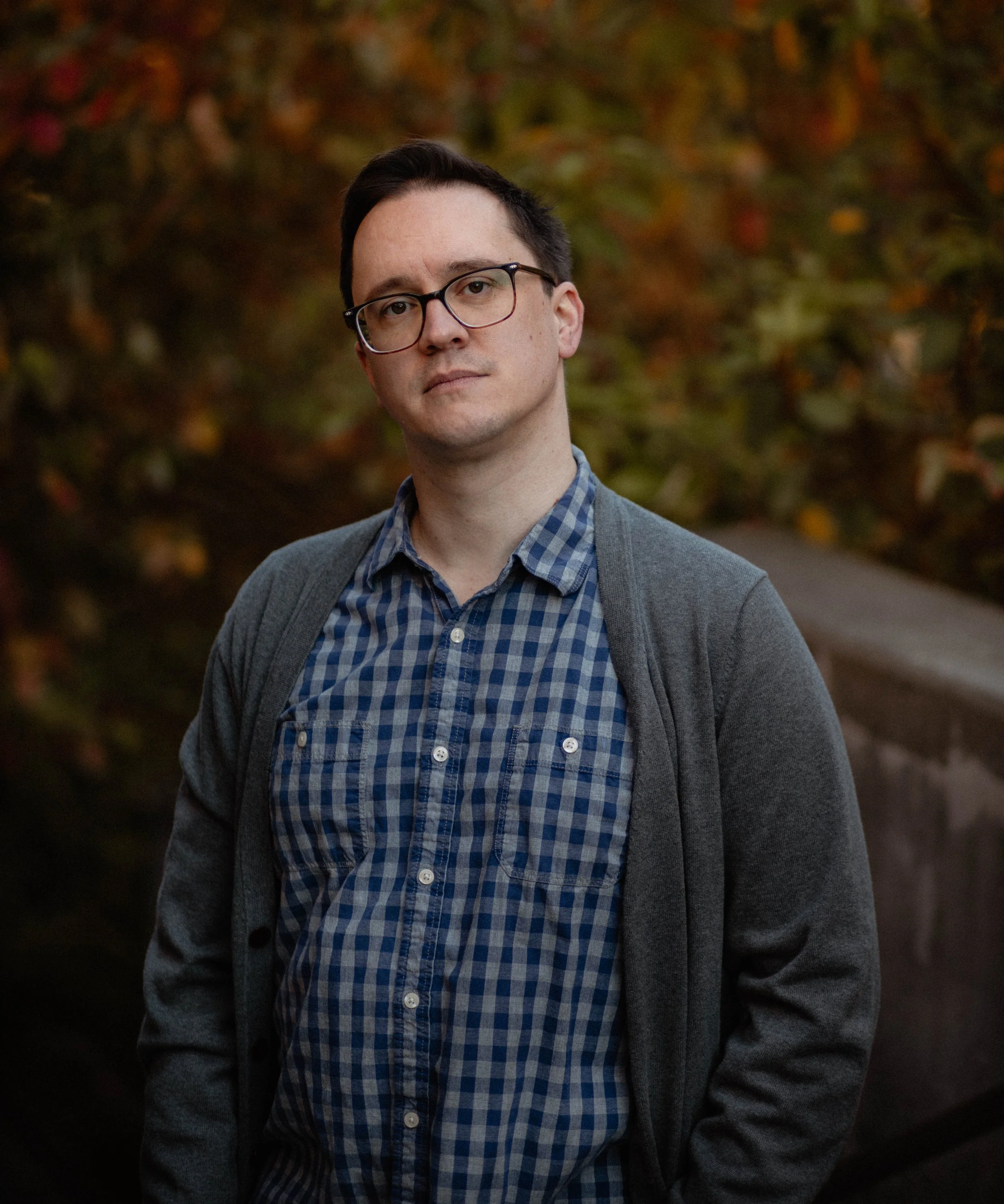 A young man with dark hair, glasses, wearing a gray cardigan over a blue plaid shirt, standing outdoors in front of autumn-colored foliage. Seattle professional head shot photography.