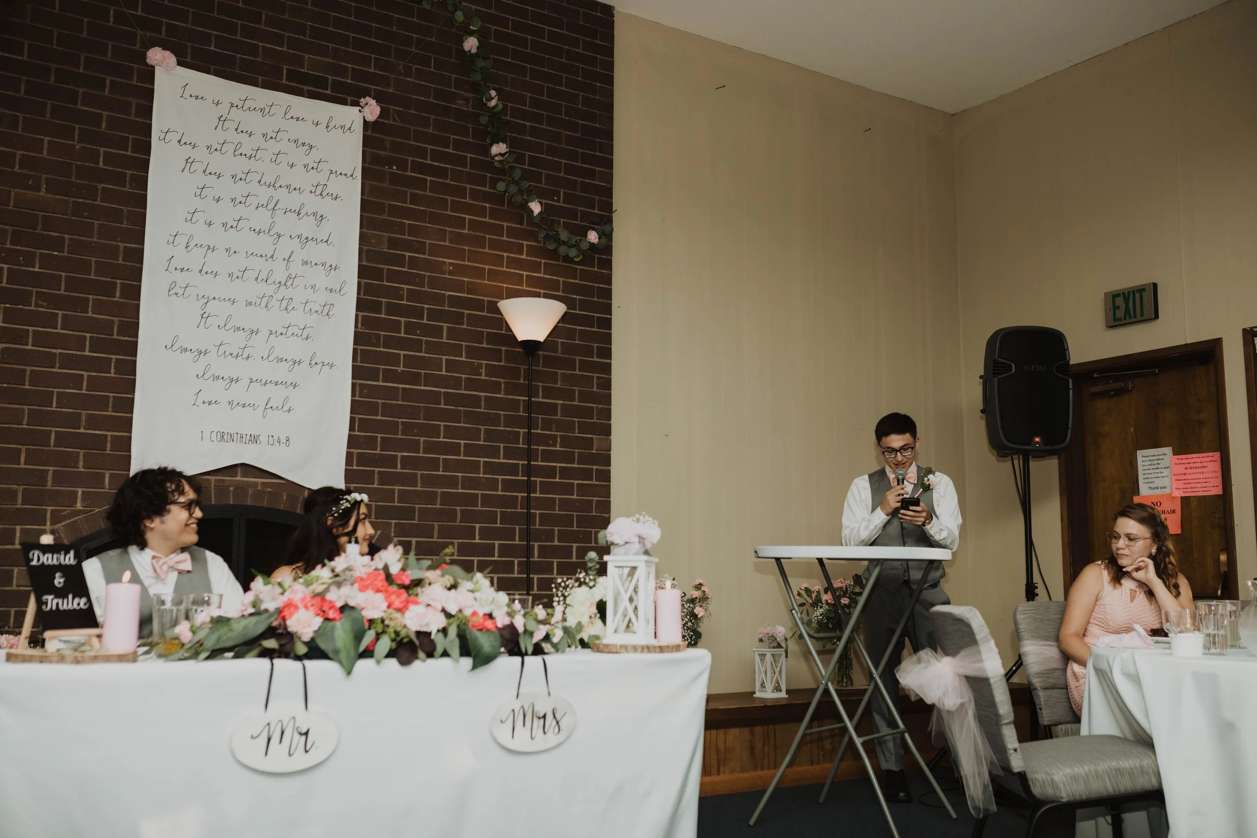 A wedding reception with the bride and groom sitting at a decorated table with floral arrangements and a pink candle, while the best man or maid of honor gives a speech at a microphone. The room has a brick wall with a large white banner displaying a