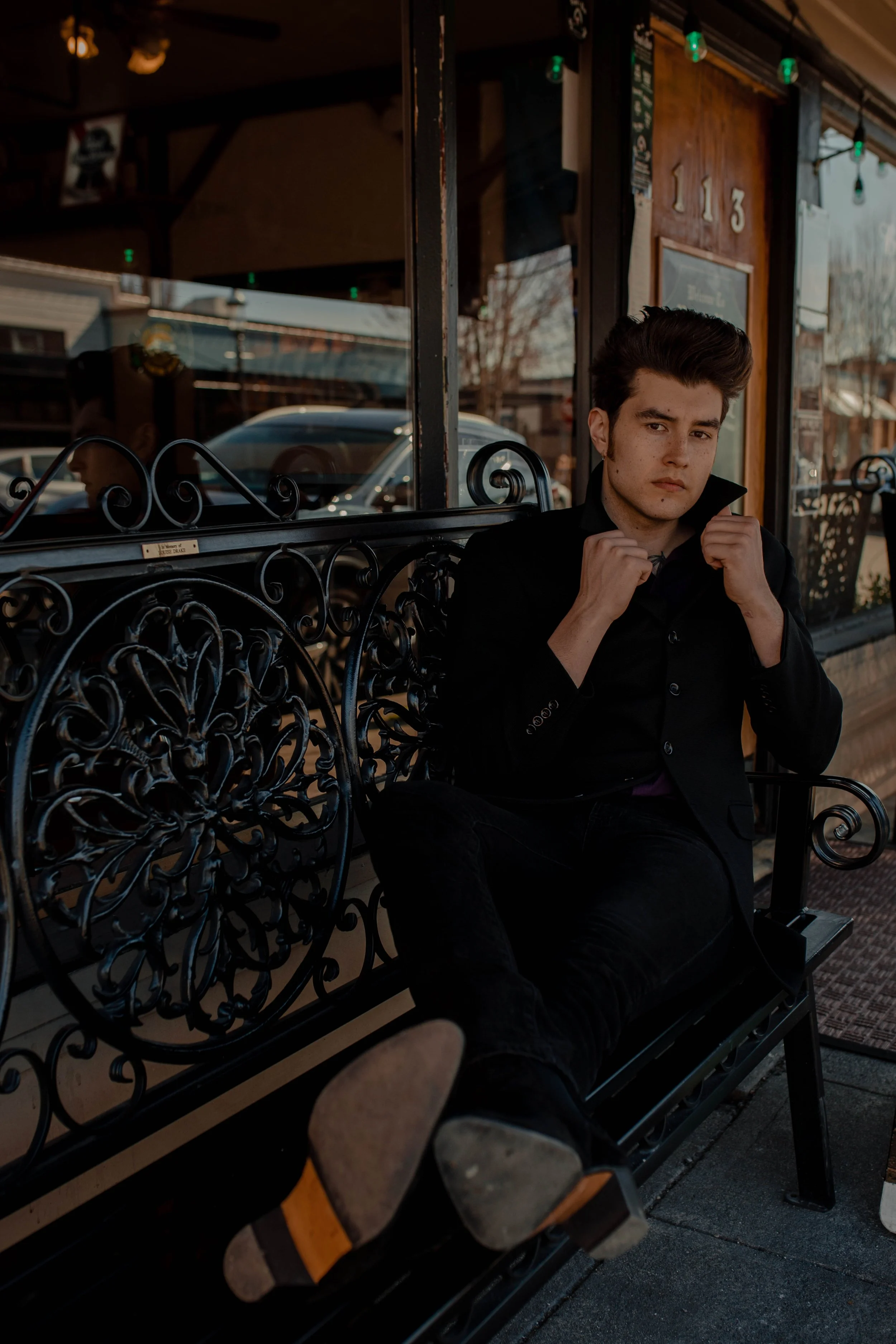 A young man with dark hair and a serious expression sitting on an ornate black metal bench outside a storefront, adjusting his collar. Seattle professional head shot photography