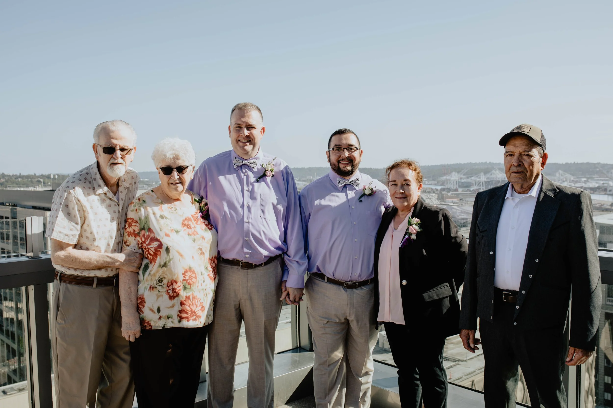 Group of seven people standing on a rooftop with a cityscape and water in the background, dressed in semi-formal clothing with some wearing boutonnieres. Seattle Municipal Courthouse wedding photography.