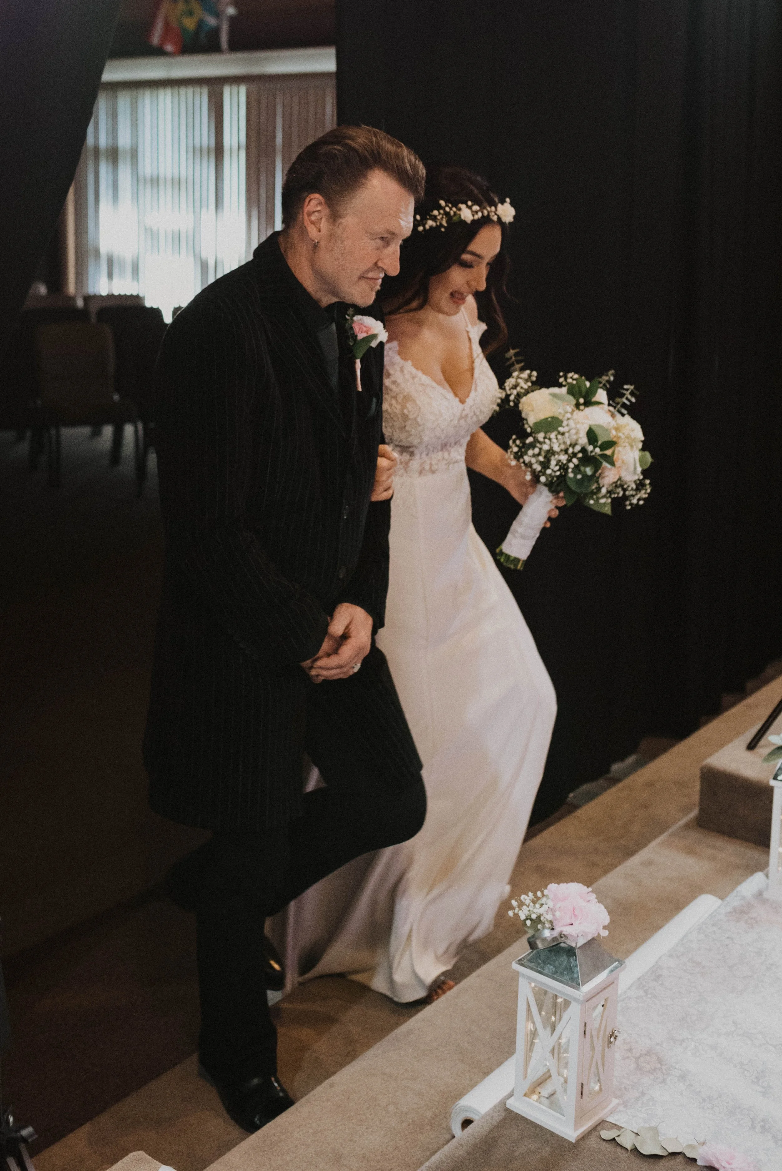 A bride in a white lace wedding dress holding a bouquet, walking arm in arm with an older man in a black pinstripe suit during a wedding ceremony. Seattle, WA wedding photography.