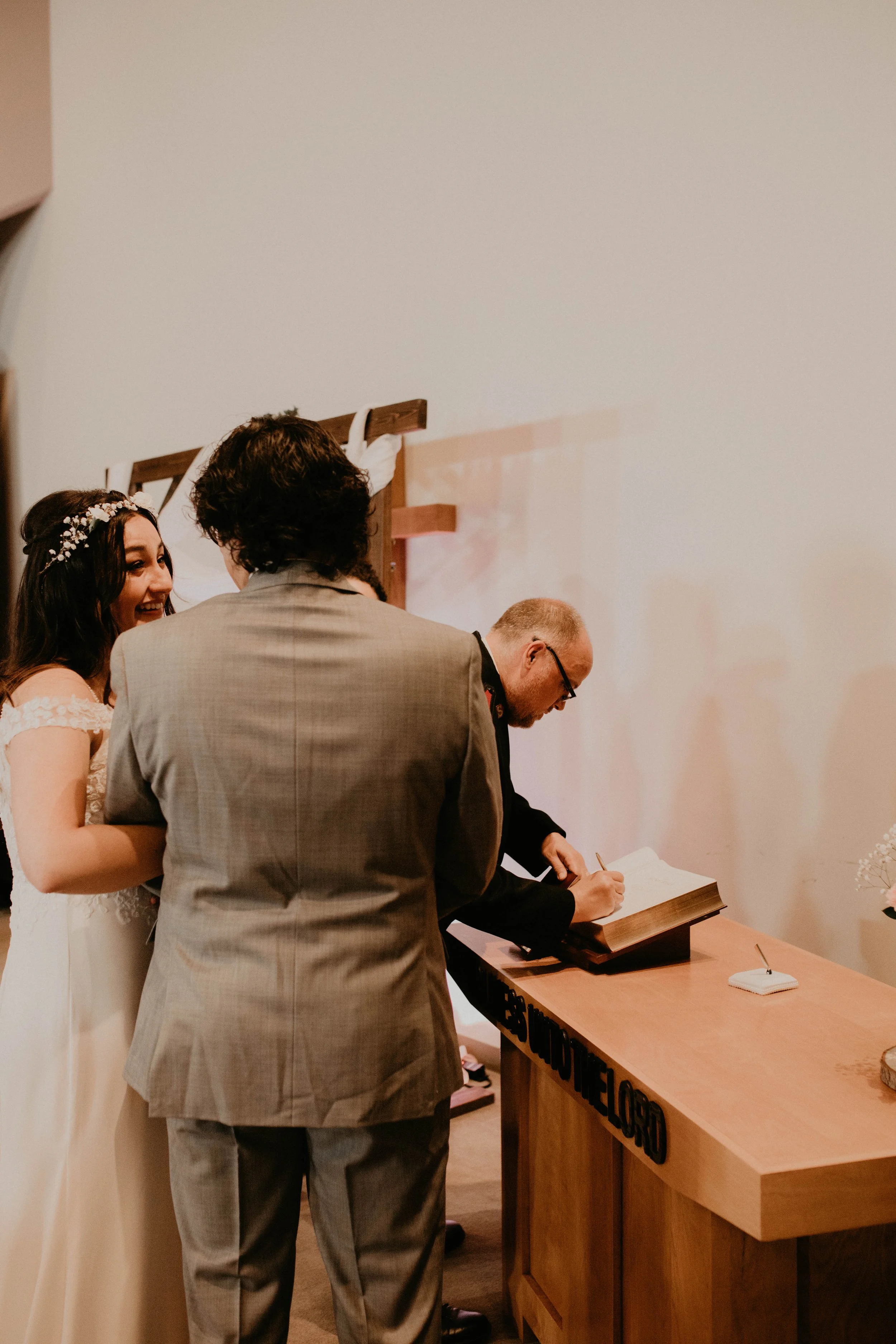 A wedding ceremony with a bride in a white dress and floral headband, a groom in a gray suit, and an officiant signing a book at a wooden altar. Seattle, WA wedding photography.