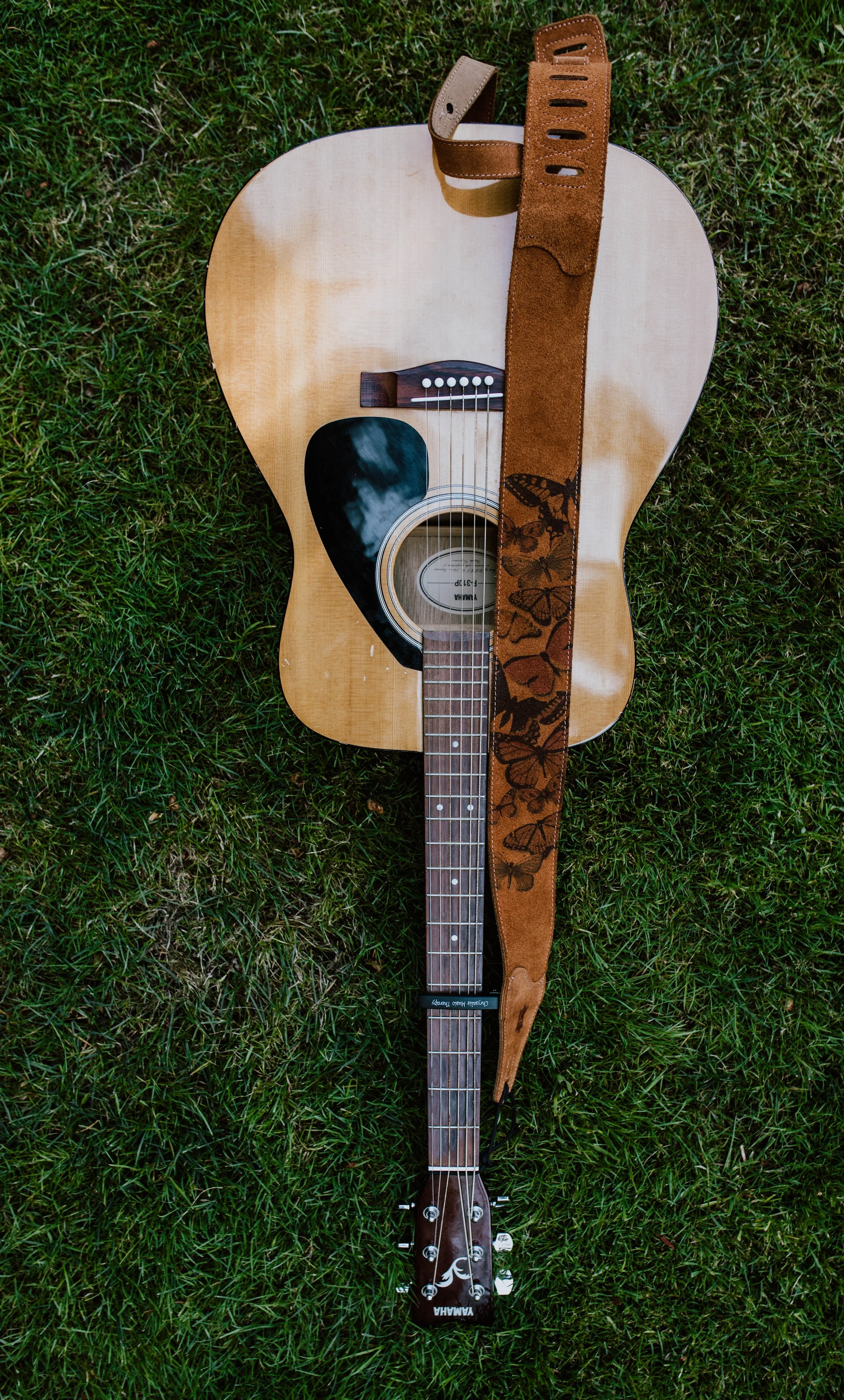 An acoustic guitar with a natural wood finish and a black pickguard, lying on green grass. The guitar has a brown strap with butterfly patterns, hanging over the body and neck. Seattle professional head shot photography