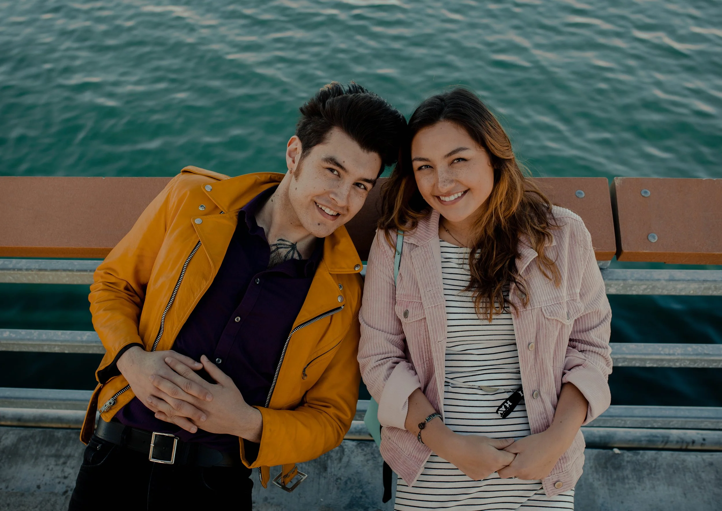 A young man with dark hair and a yellow jacket, and a young woman with long brown hair and a pink jacket, smiling and standing near water on a pier. Seattle professional head shot photography
