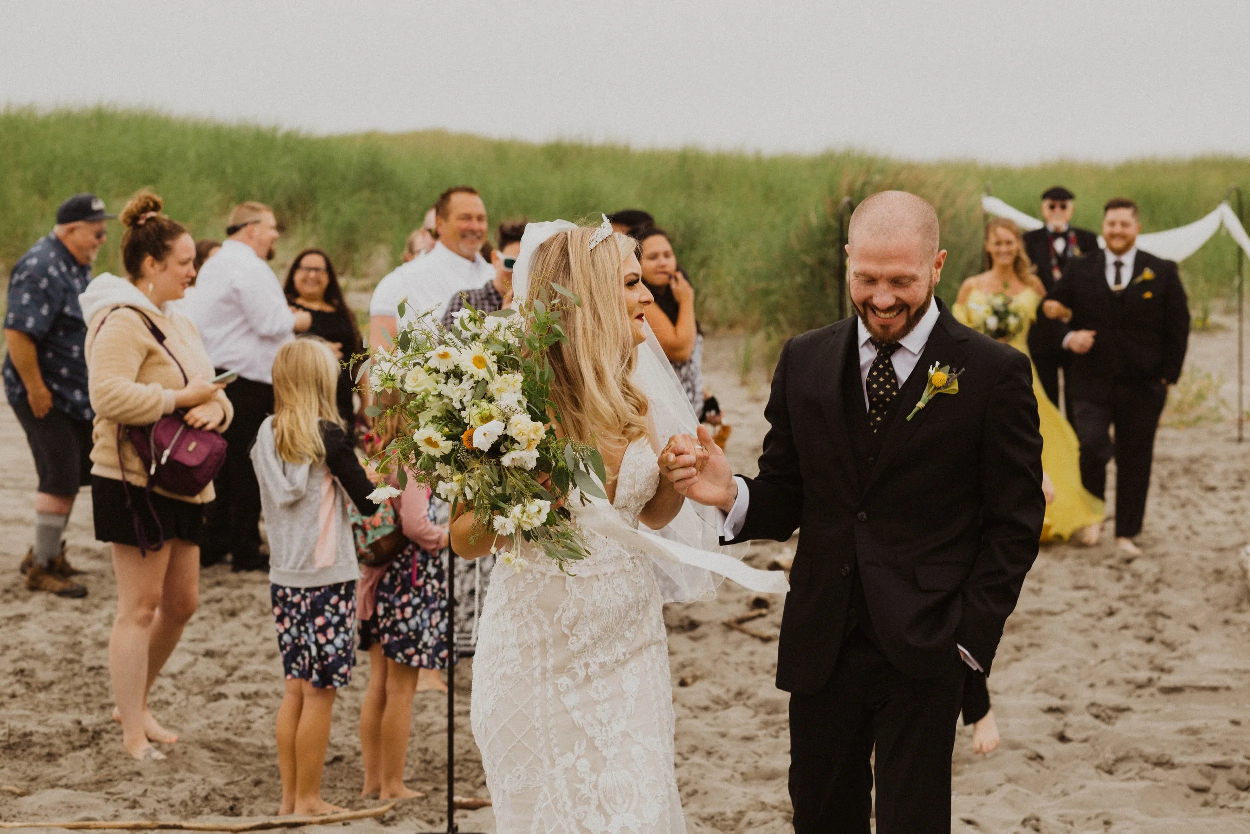 A couple in wedding attire smiling and holding hands on a sandy beach, surrounded by friends and family during a wedding celebration. Long Beach, WA wedding photography.