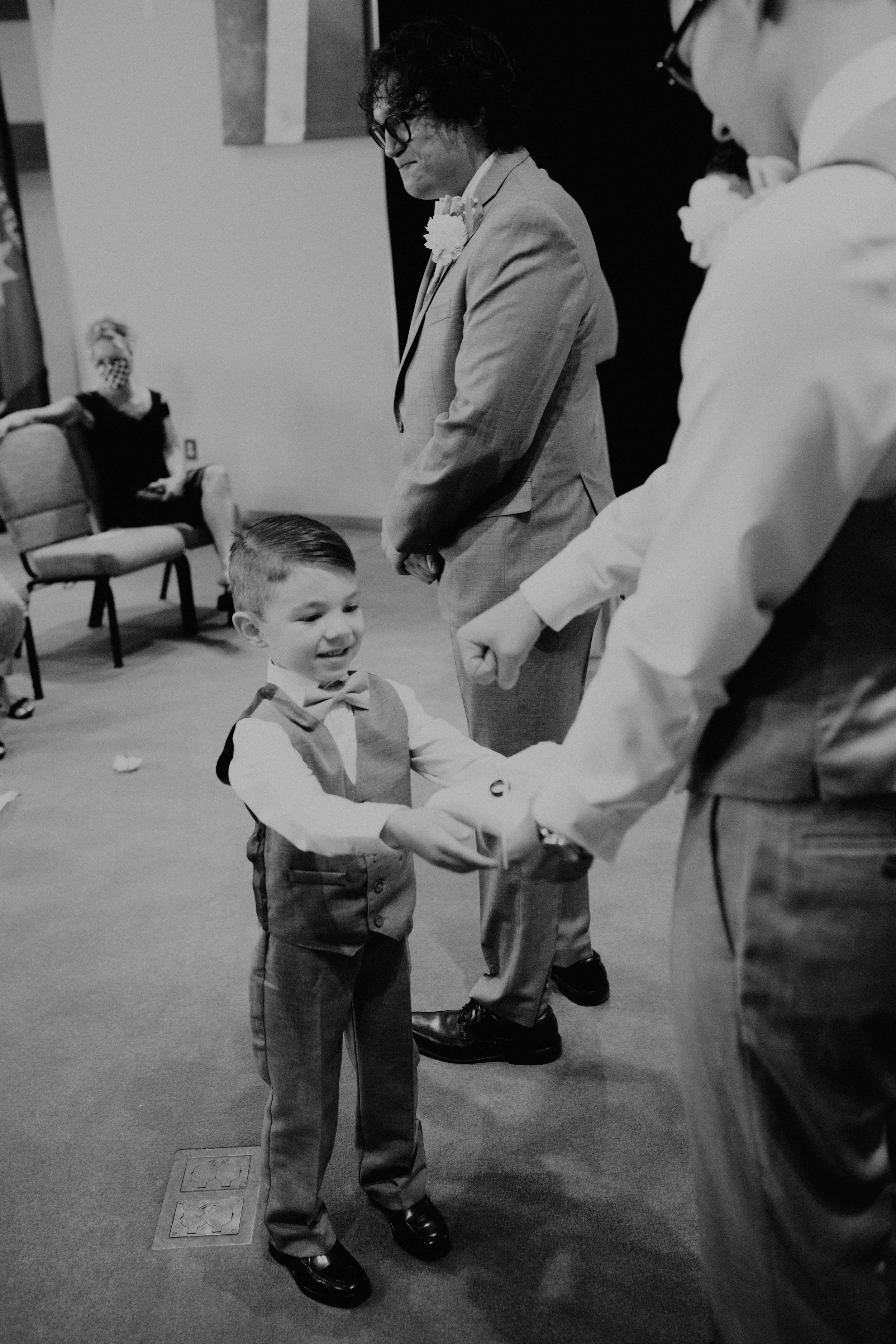 A young boy in formal attire holding a gift, smiling, in a room with adults. An adult man in a suit stands behind him, and other people are sitting in the background. Seattle, WA wedding photography.