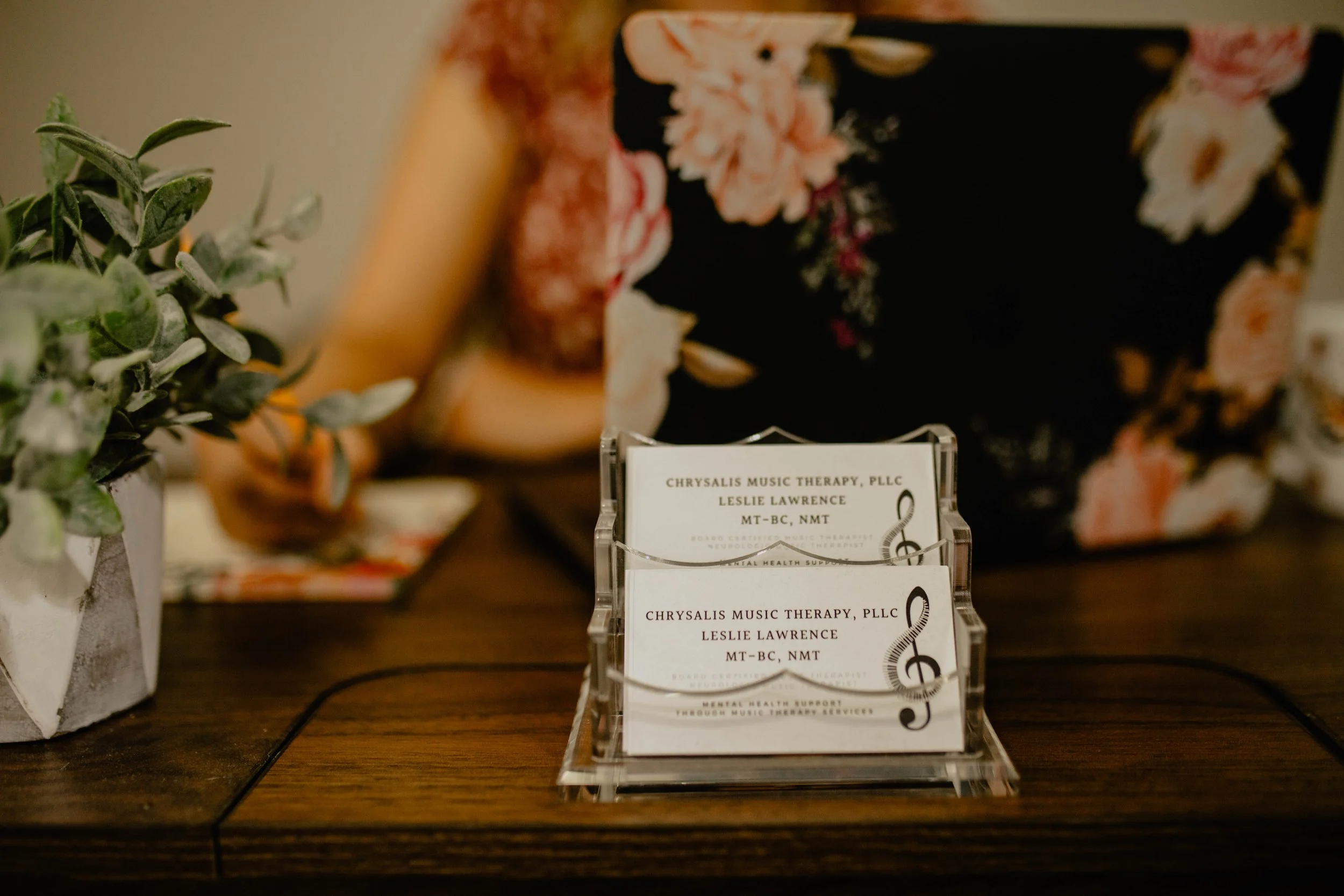 Business cards for Chrysalis Music Therapy, PLLC, Leslie Lawrence, placed in a clear card holder on a wooden table, with a potted plant on the left and a woman using a laptop in the background. Seattle professional head shot photography