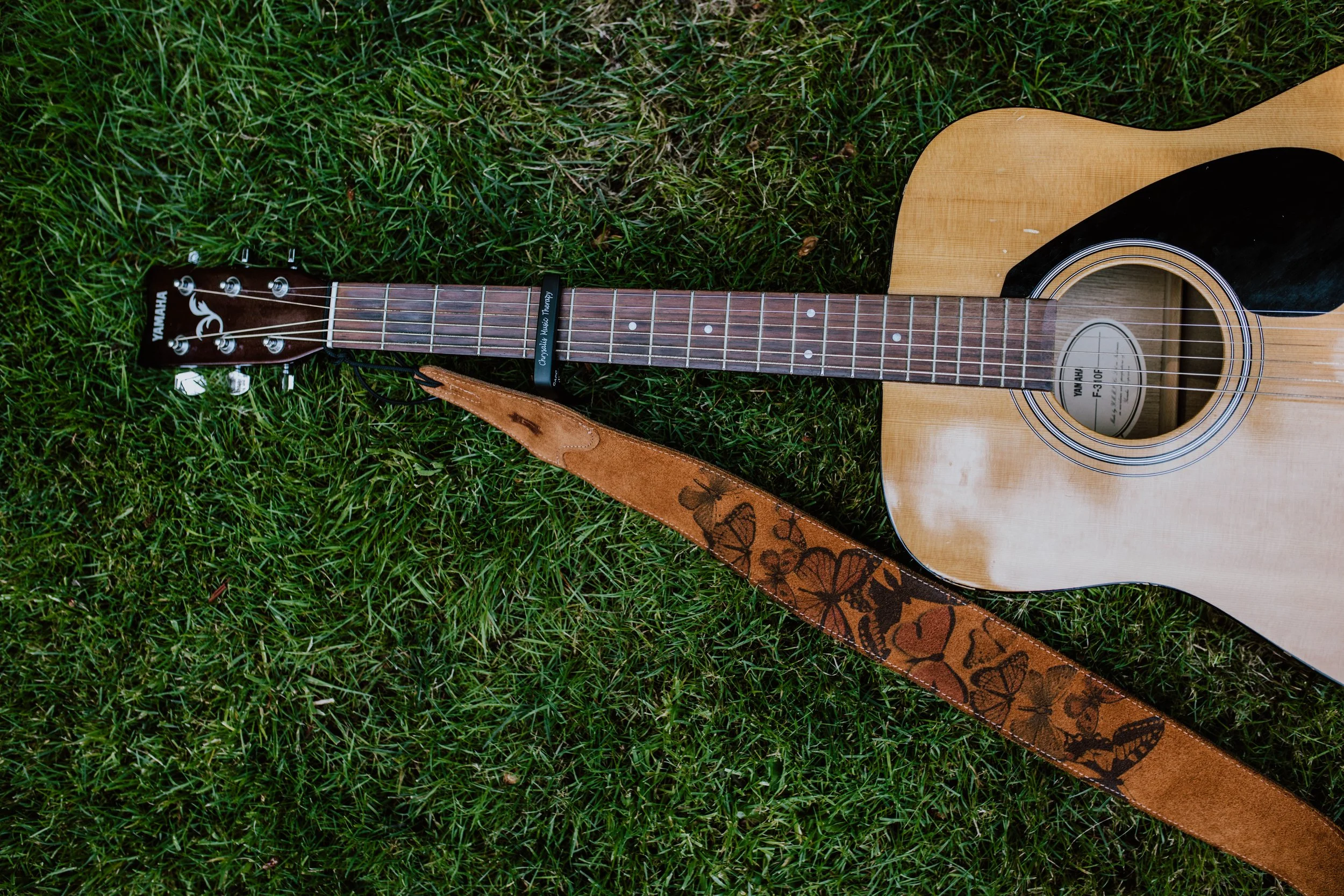An acoustic guitar lying on green grass, with a strap featuring butterfly and heart designs. Seattle professional head shot photography