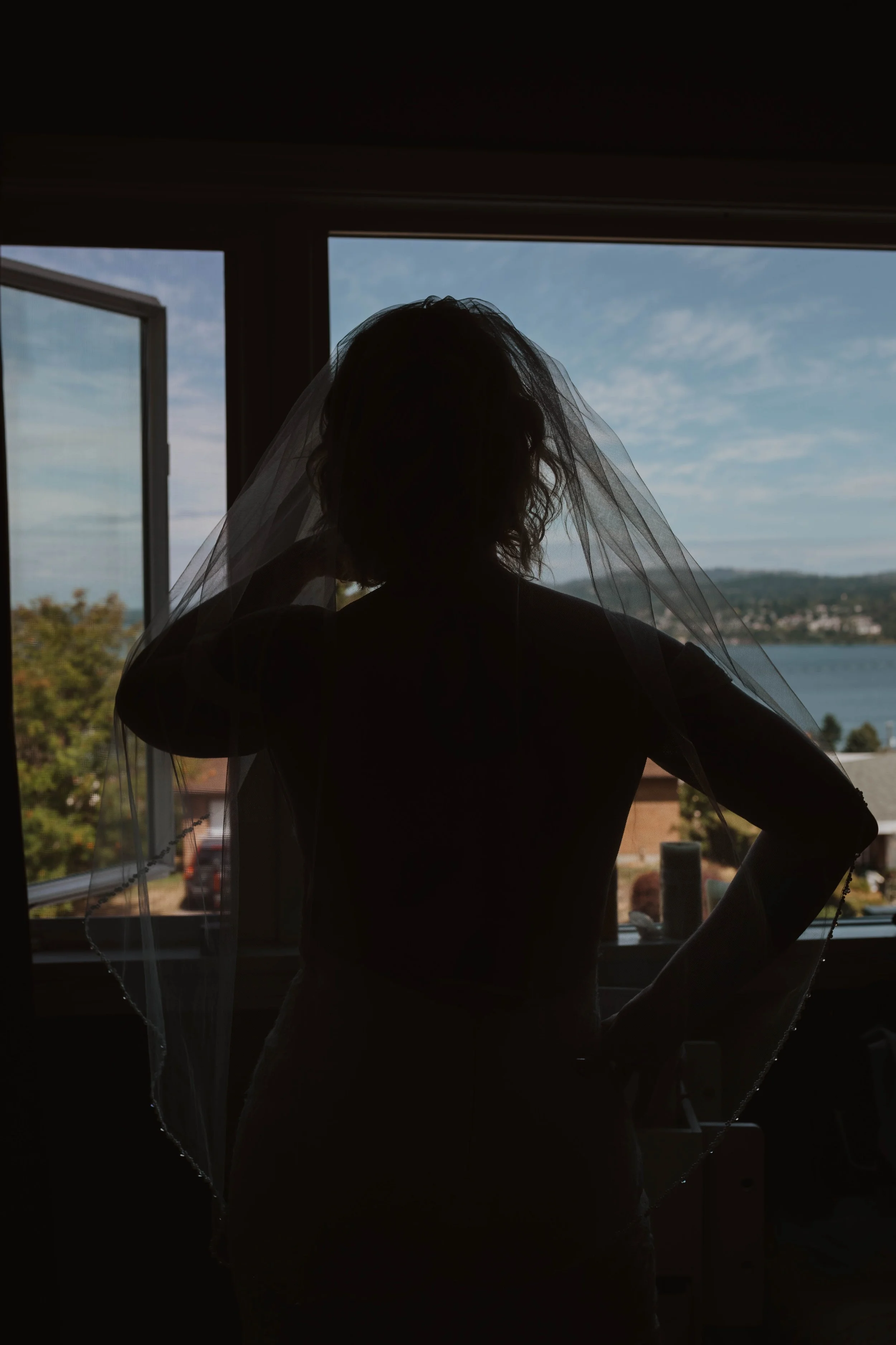 A woman wearing a veil standing in front of a window with a view of a lake, trees, and houses outside during daytime. Seattle, WA wedding photography.
