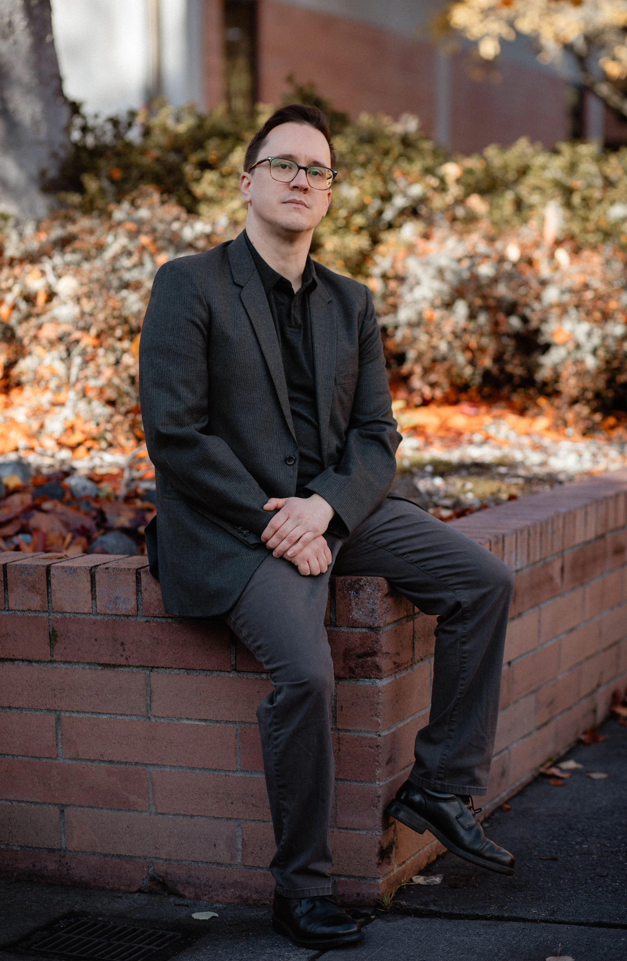 A man in a dark suit sits on a brick ledge outdoors, with autumn leaves and bushes in the background. Seattle professional head shot photography
