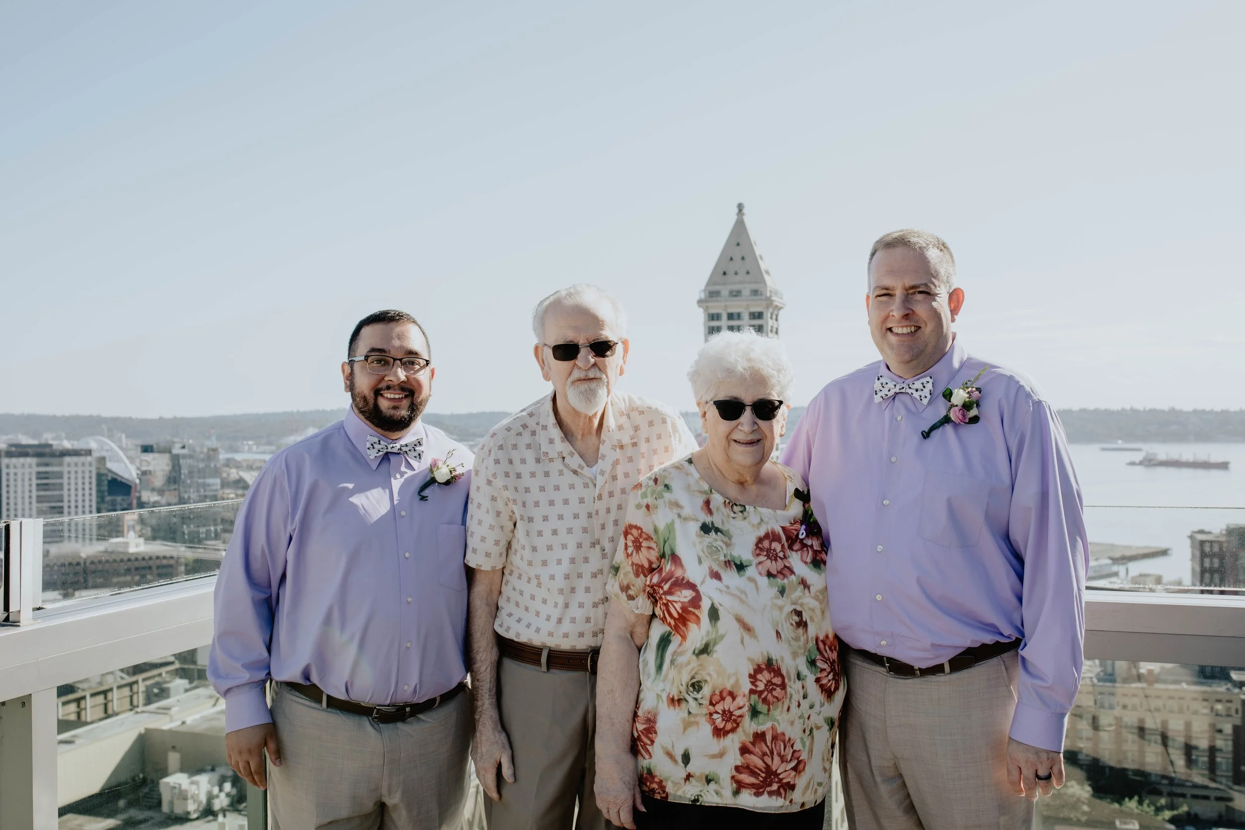 Group of five people standing on a rooftop with city skyline and water in the background, smiling, dressed for a formal occasion, two men wearing light purple shirts and bow ties, an elderly man and woman in floral outfits.